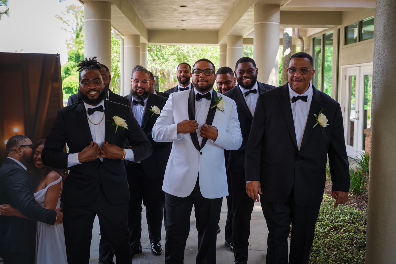 Groom in white tuxedo jacket poses with groomsmen in black tuxedos and boutonnieres at outdoor wedding venue.