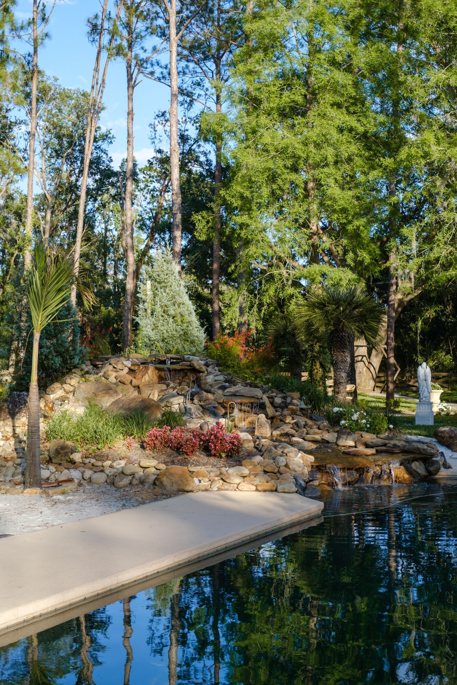 Tranquil swimming pool surrounded by natural landscaping with stone waterfall feature and tall trees reflecting in blue water.