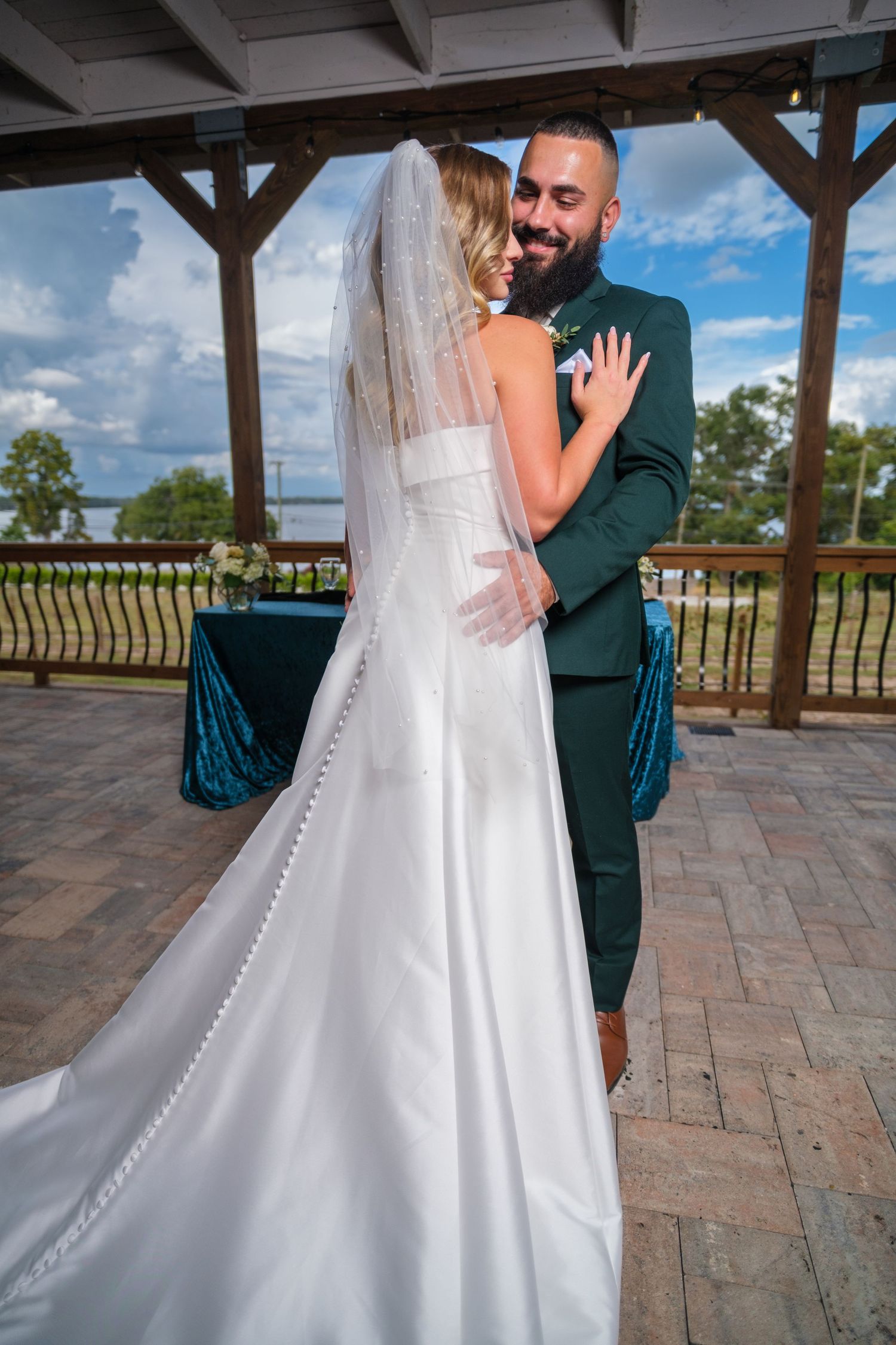 A bride in a flowing white gown and veil shares a tender moment with her partner on a paved outdoor terrace.