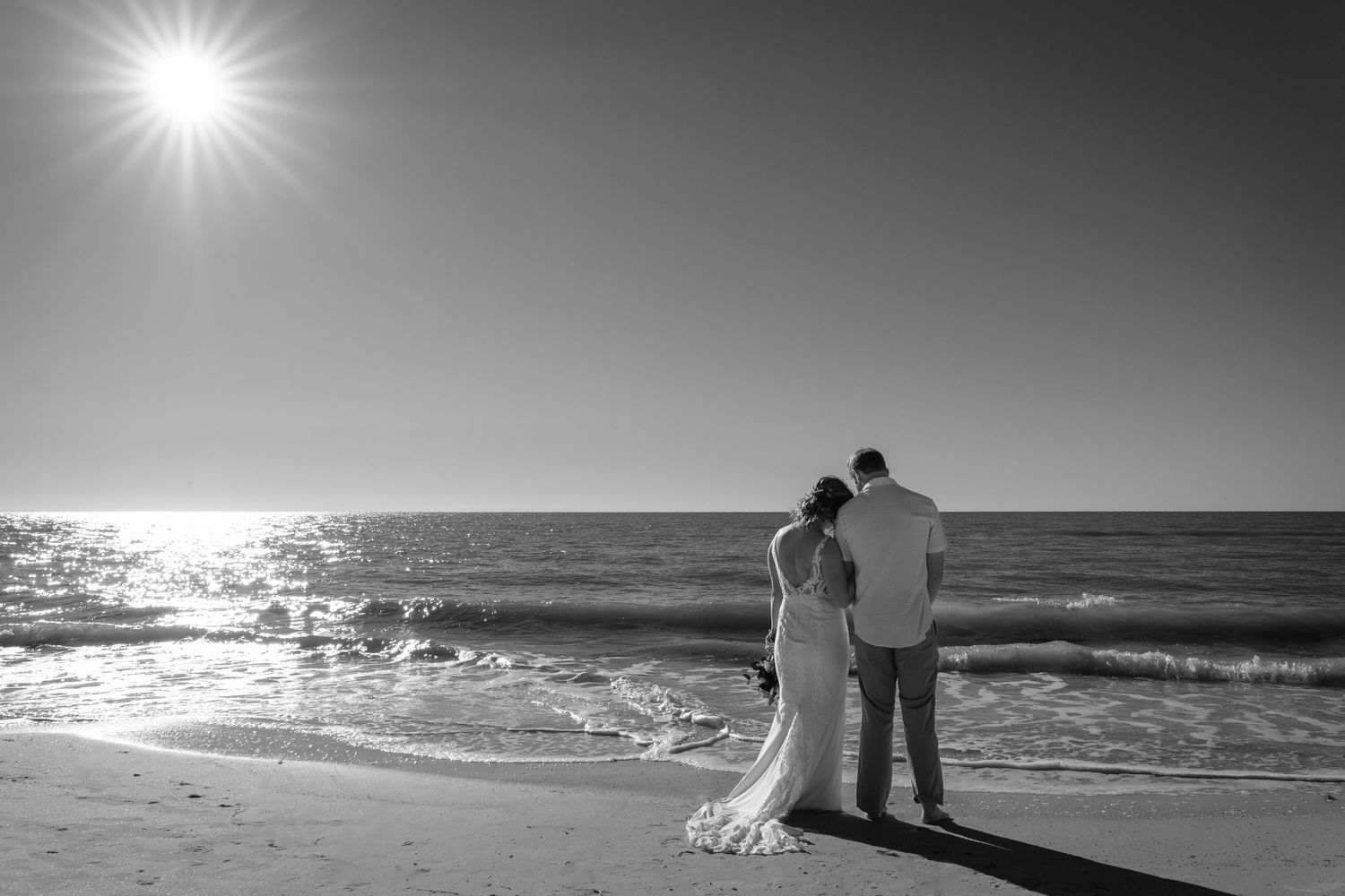 A couple walks along a scenic beach at sunset with bright sunlight creating silhouettes against the ocean waves.
