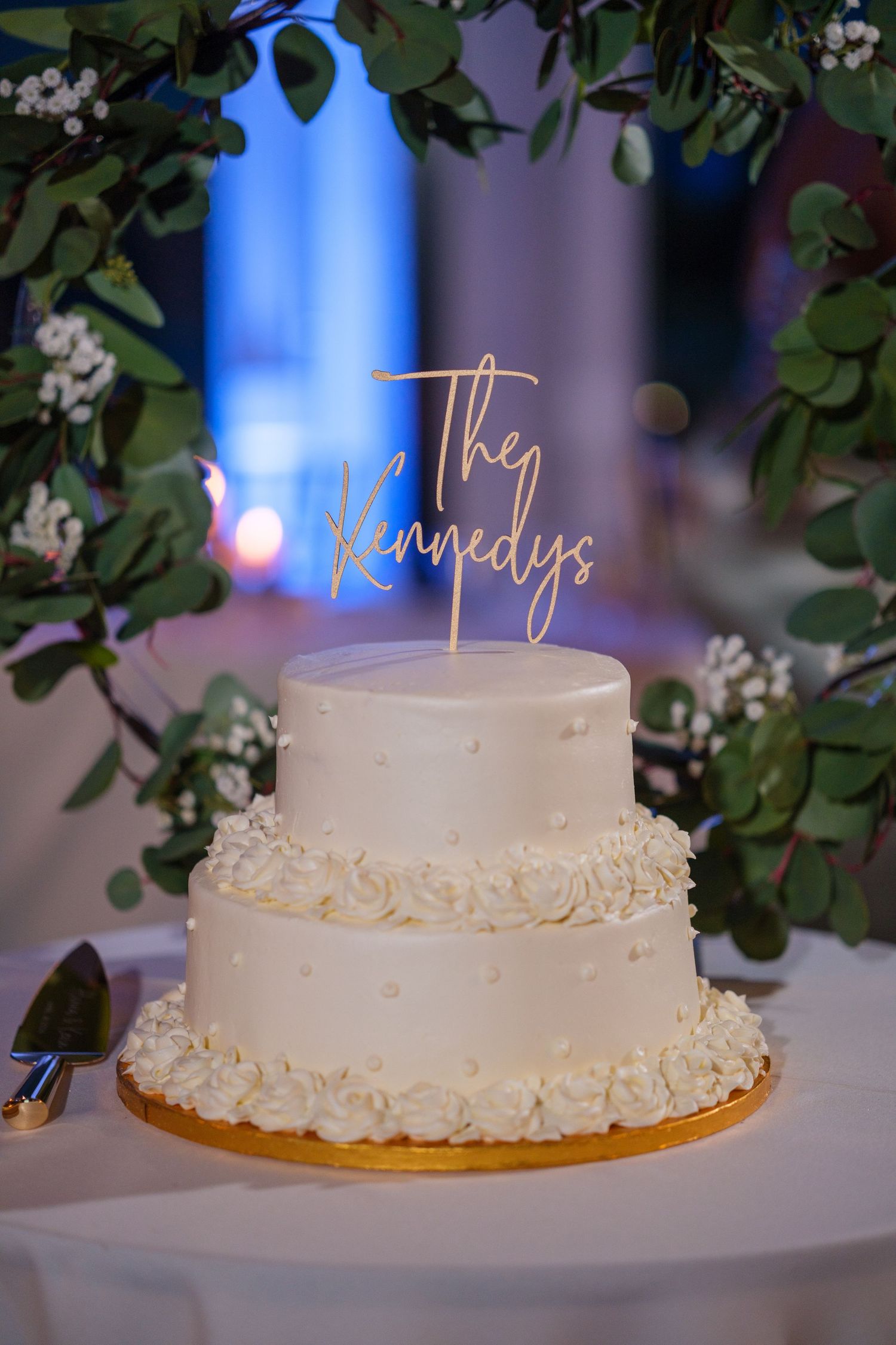 Two-tier white wedding cake with rosette details and gold The Kennedys cake topper, surrounded by greenery.