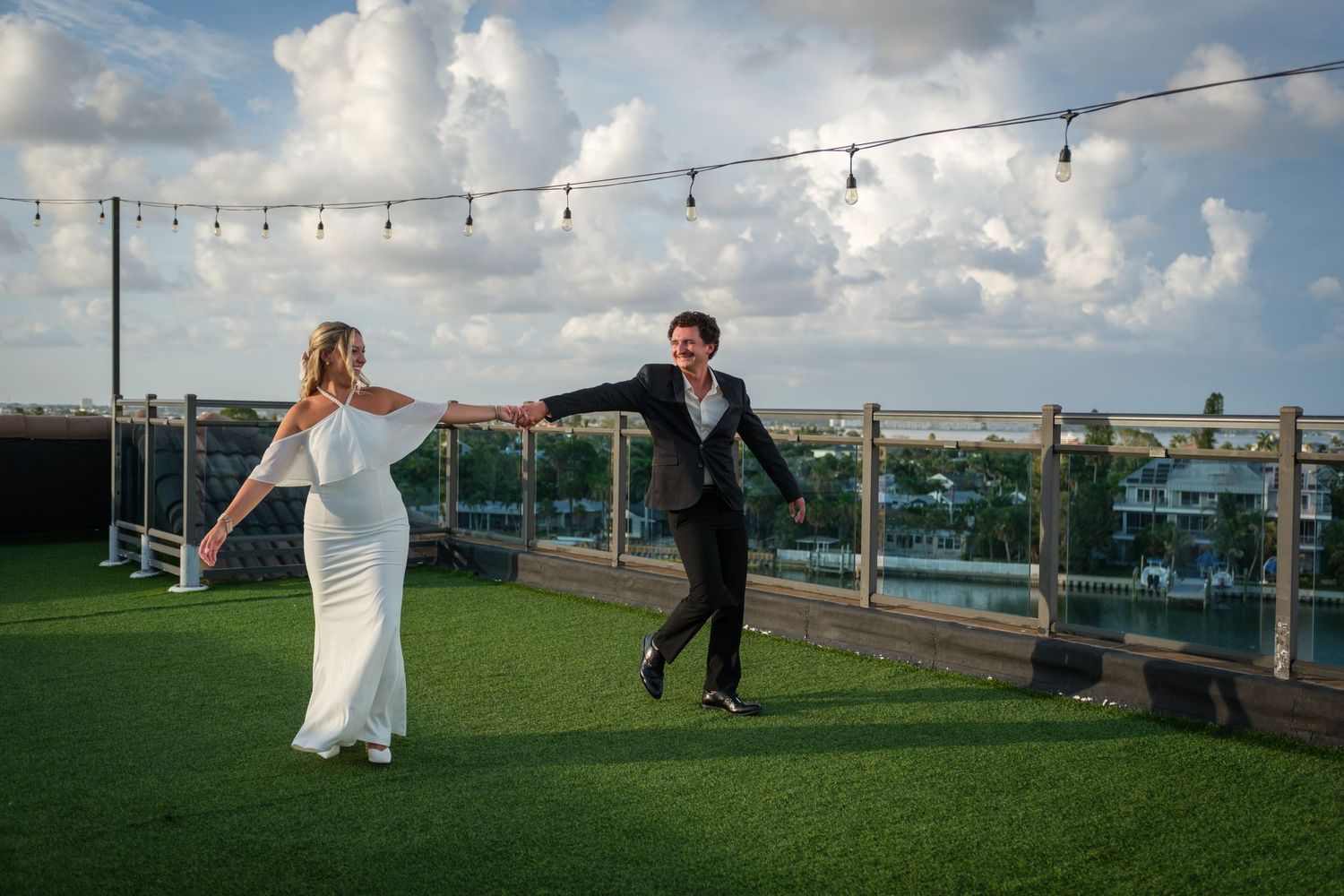 Bride and groom dancing joyfully on a rooftop venue with string lights, lush greenery, and a scenic waterfront backdrop.