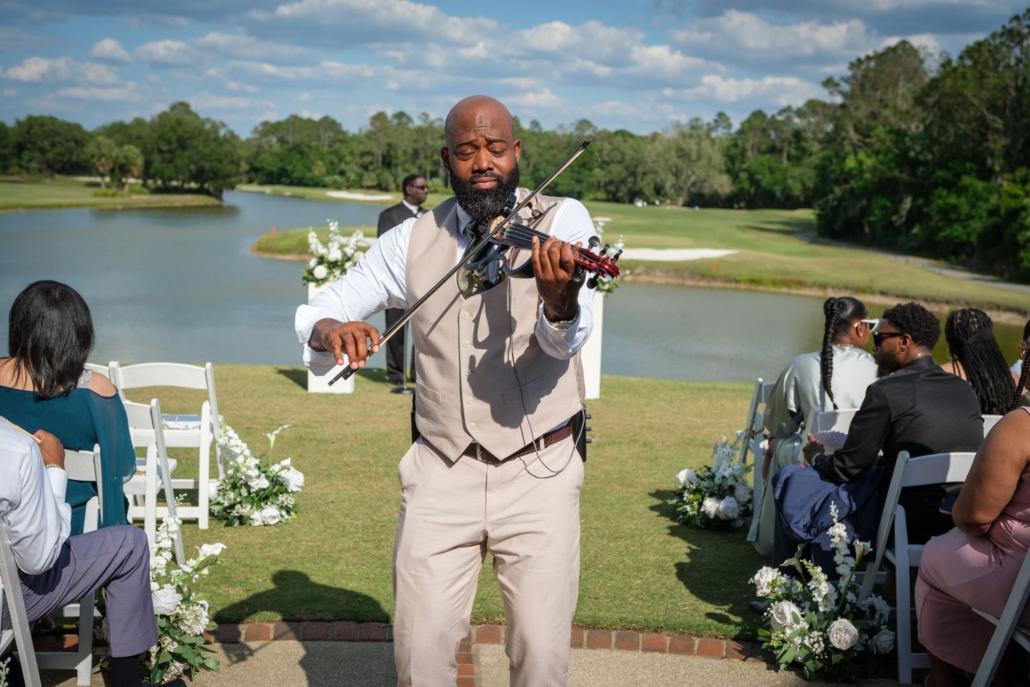 Violinist performing at outdoor wedding ceremony by a lake, wearing beige vest and dress shirt.