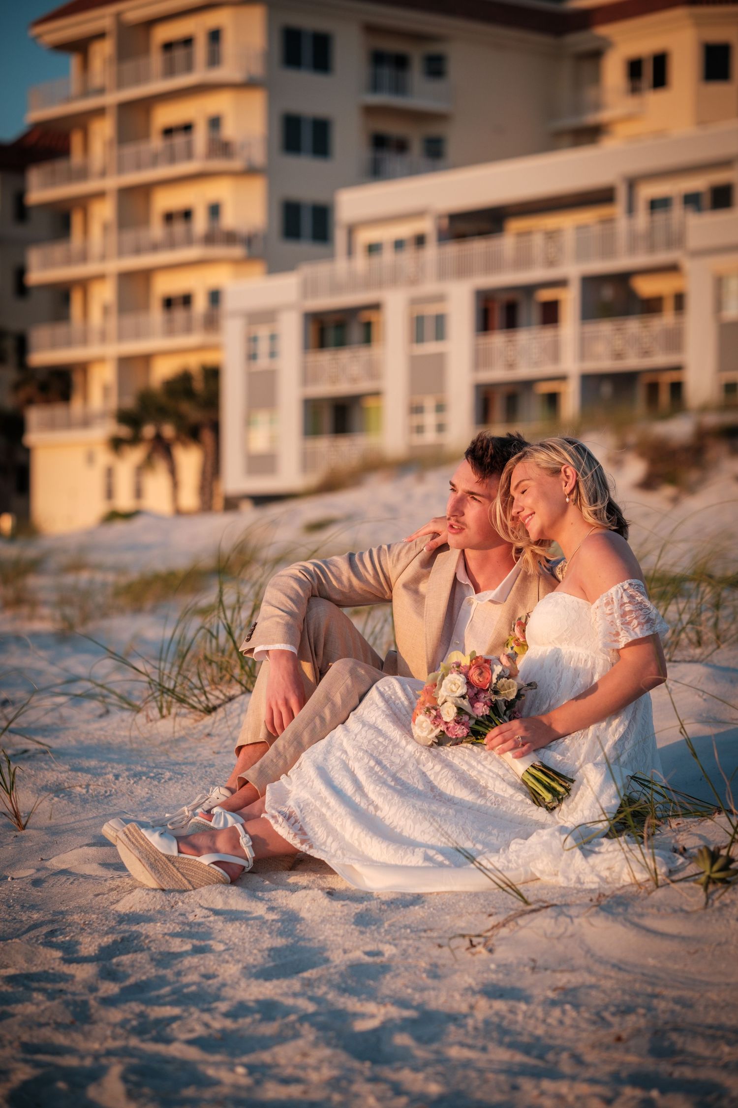 Bride and groom sitting on white sand beach at sunset, with a bouquet and resort building in the background.