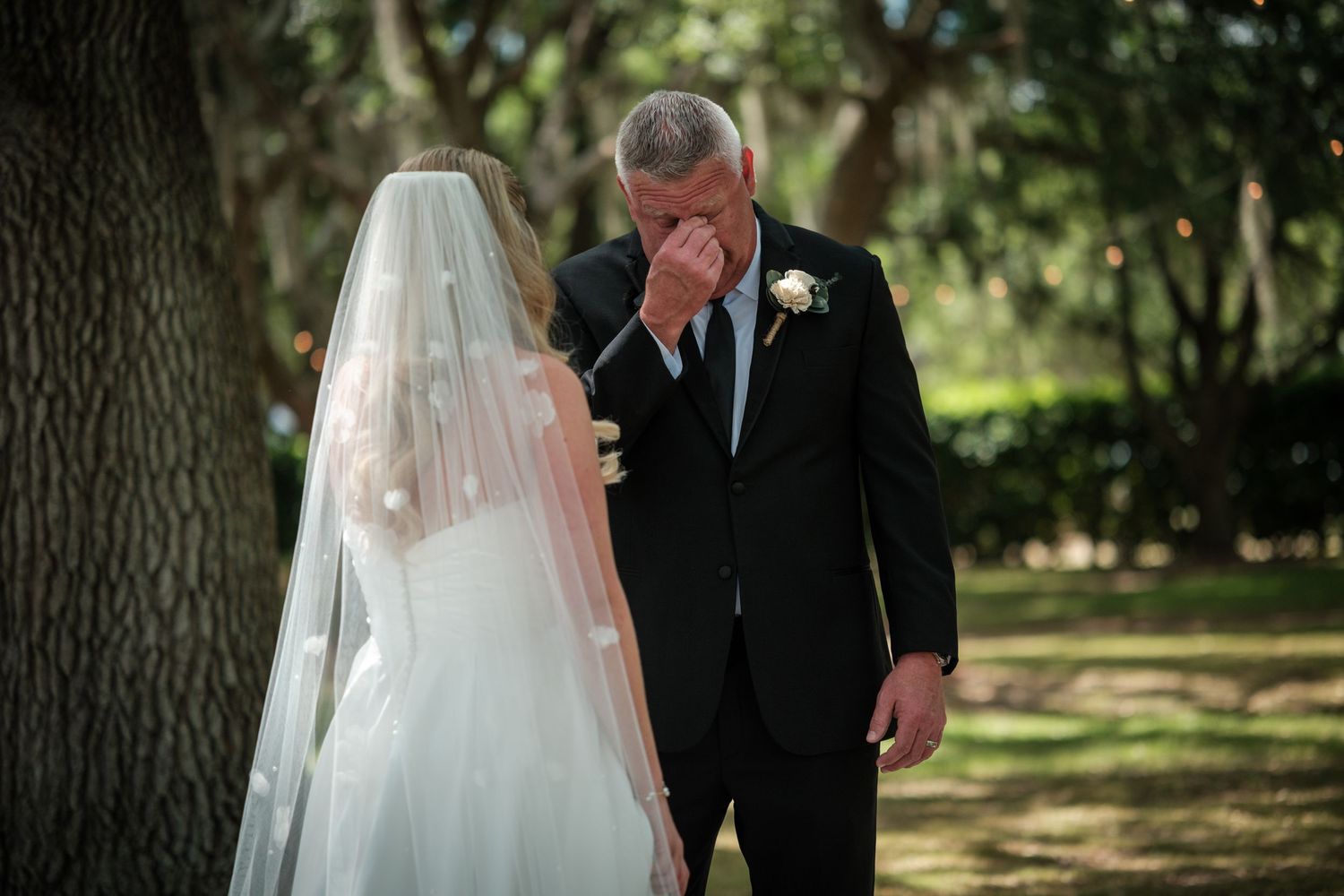 Emotional father crying during first look at bride daughter in wedding veil outdoors under trees.