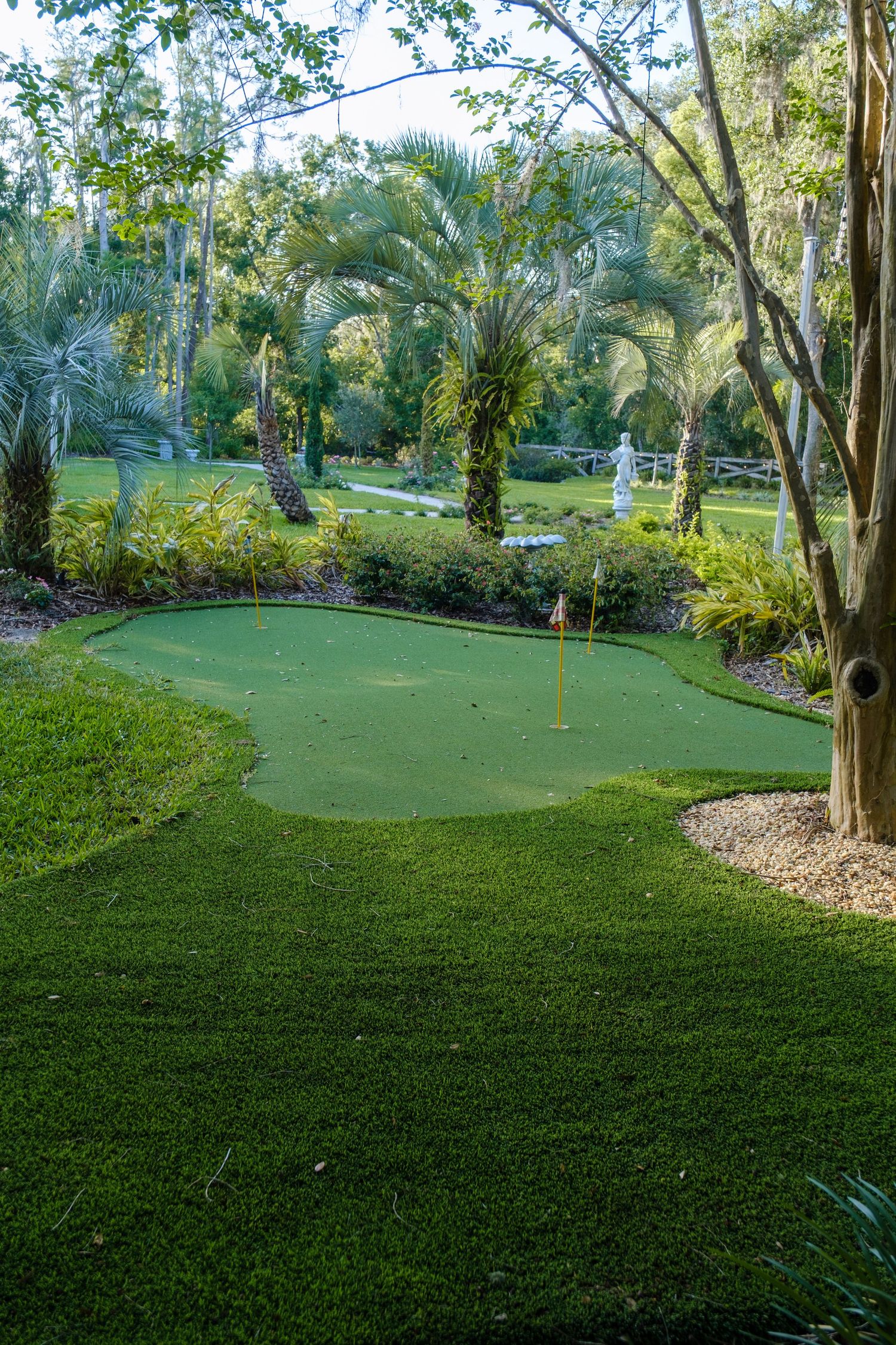 A small putting green surrounded by tropical landscaping with palm trees and lush gardens in a backyard setting.