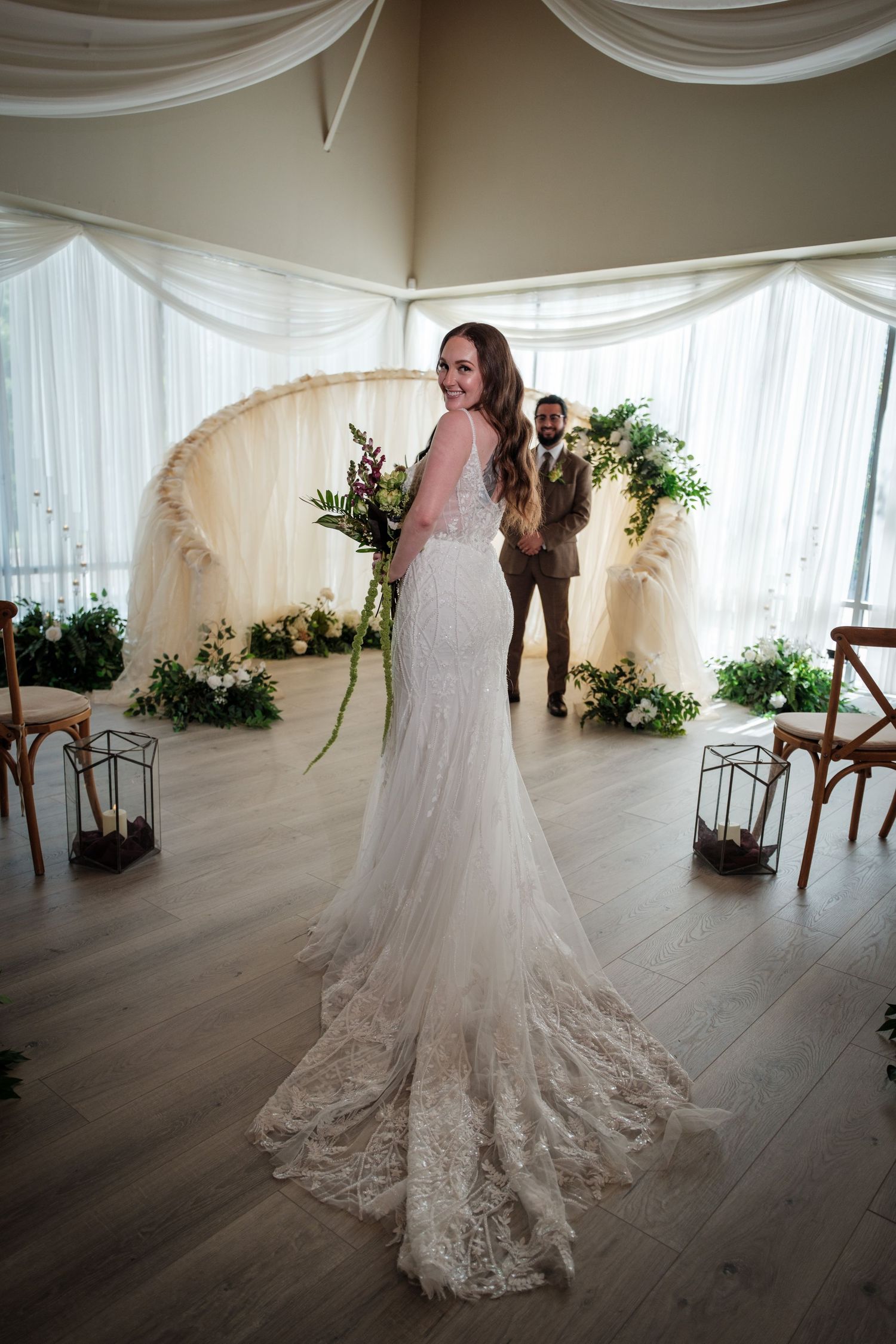 Bride in lace wedding gown with train stands before floral arch, groom in tan suit visible in background at indoor ceremony.