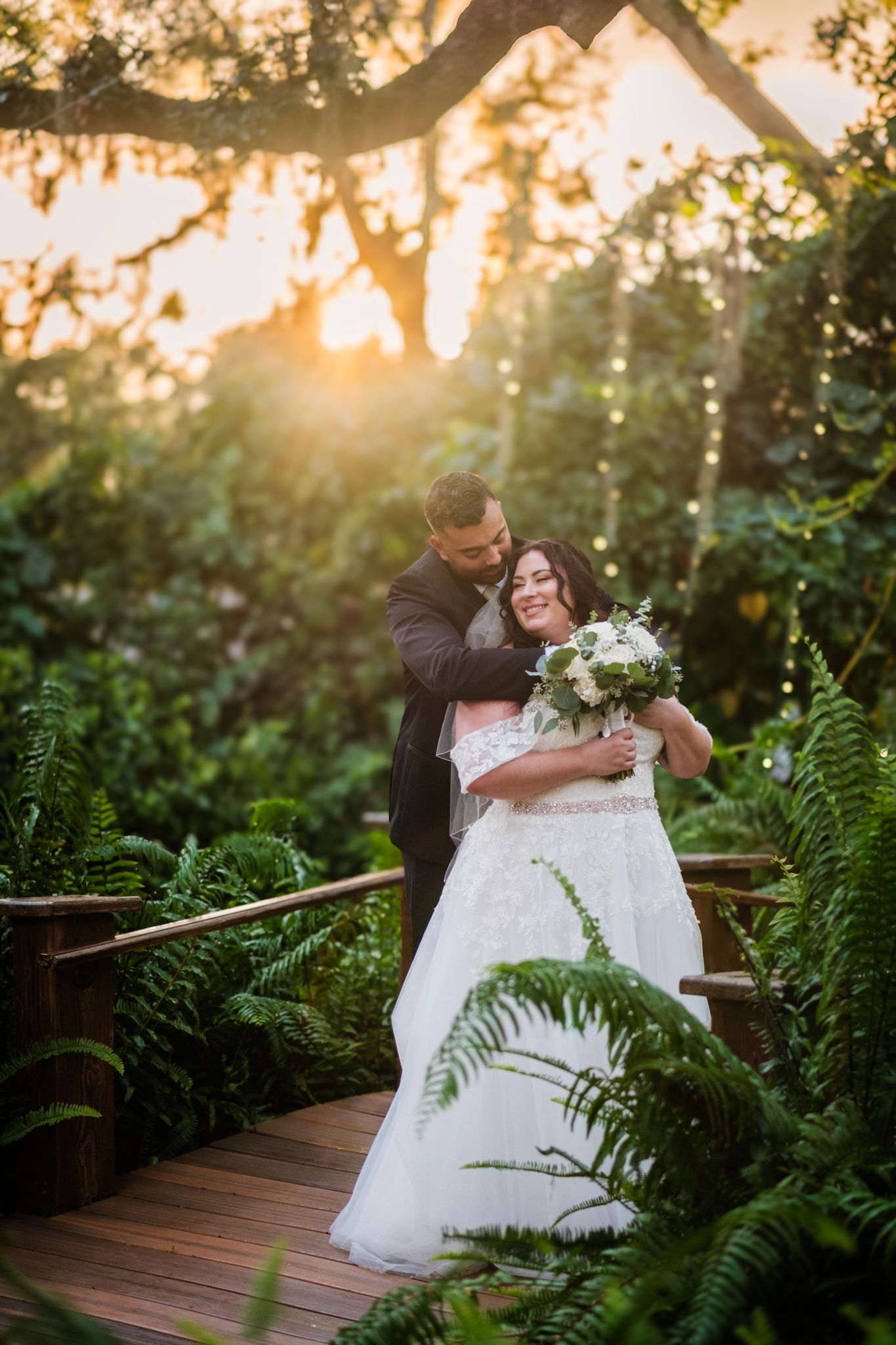 A romantic sunset wedding moment captured on a wooden boardwalk surrounded by lush ferns and greenery.