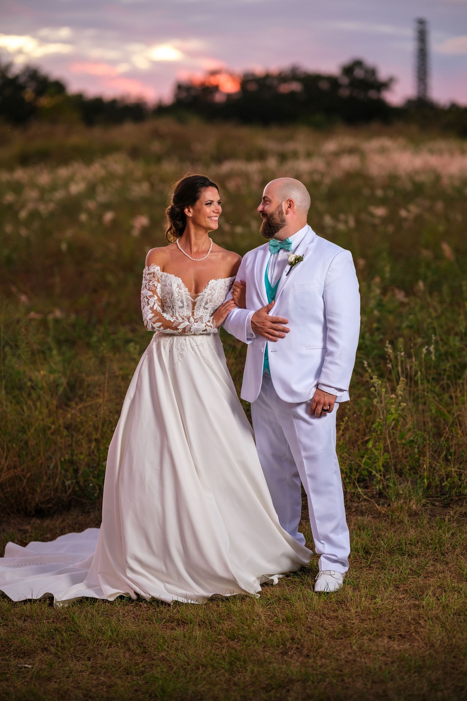 A couple in wedding attire dances together in a field at sunset with the bride wearing a flowing white dress.