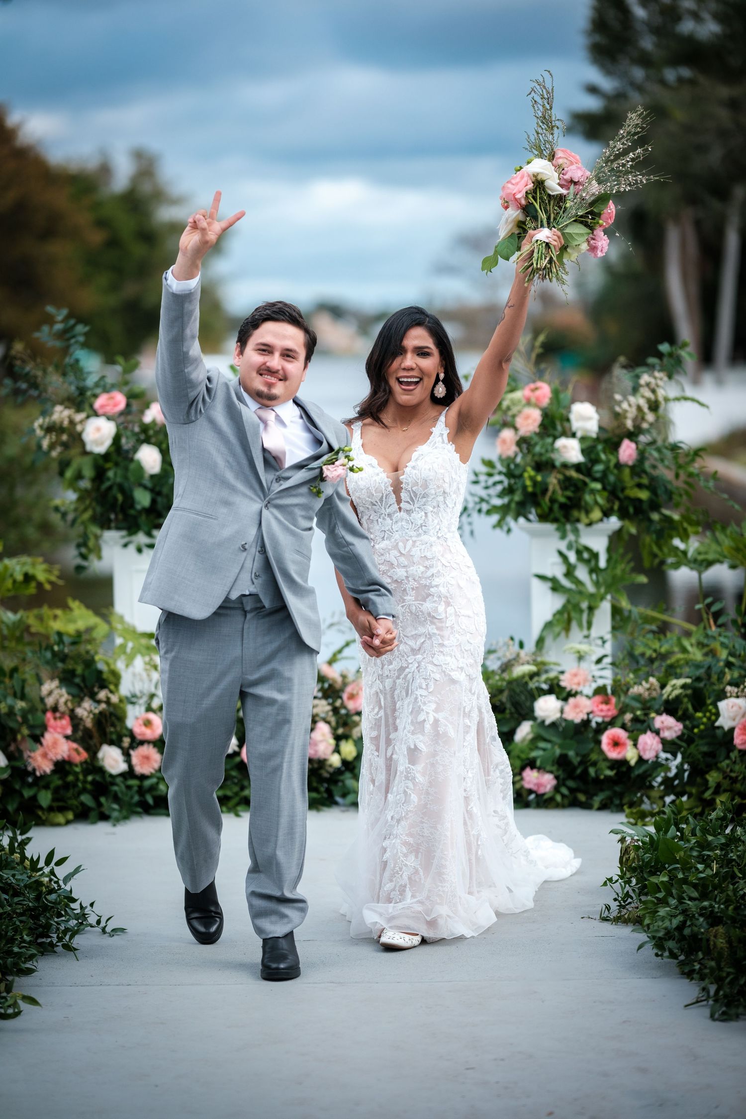 A joyful couple in wedding attire strikes fun poses along a garden path lined with pink roses and greenery.