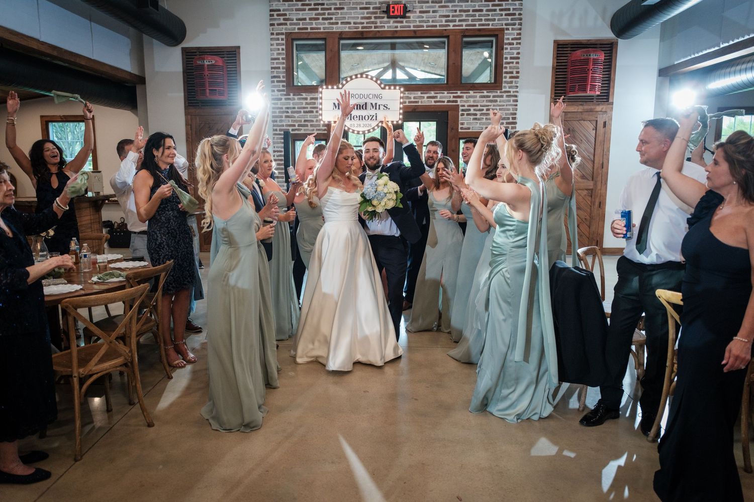 Bride and groom walk through cheering bridesmaids in sage green dresses at elegant indoor wedding reception.