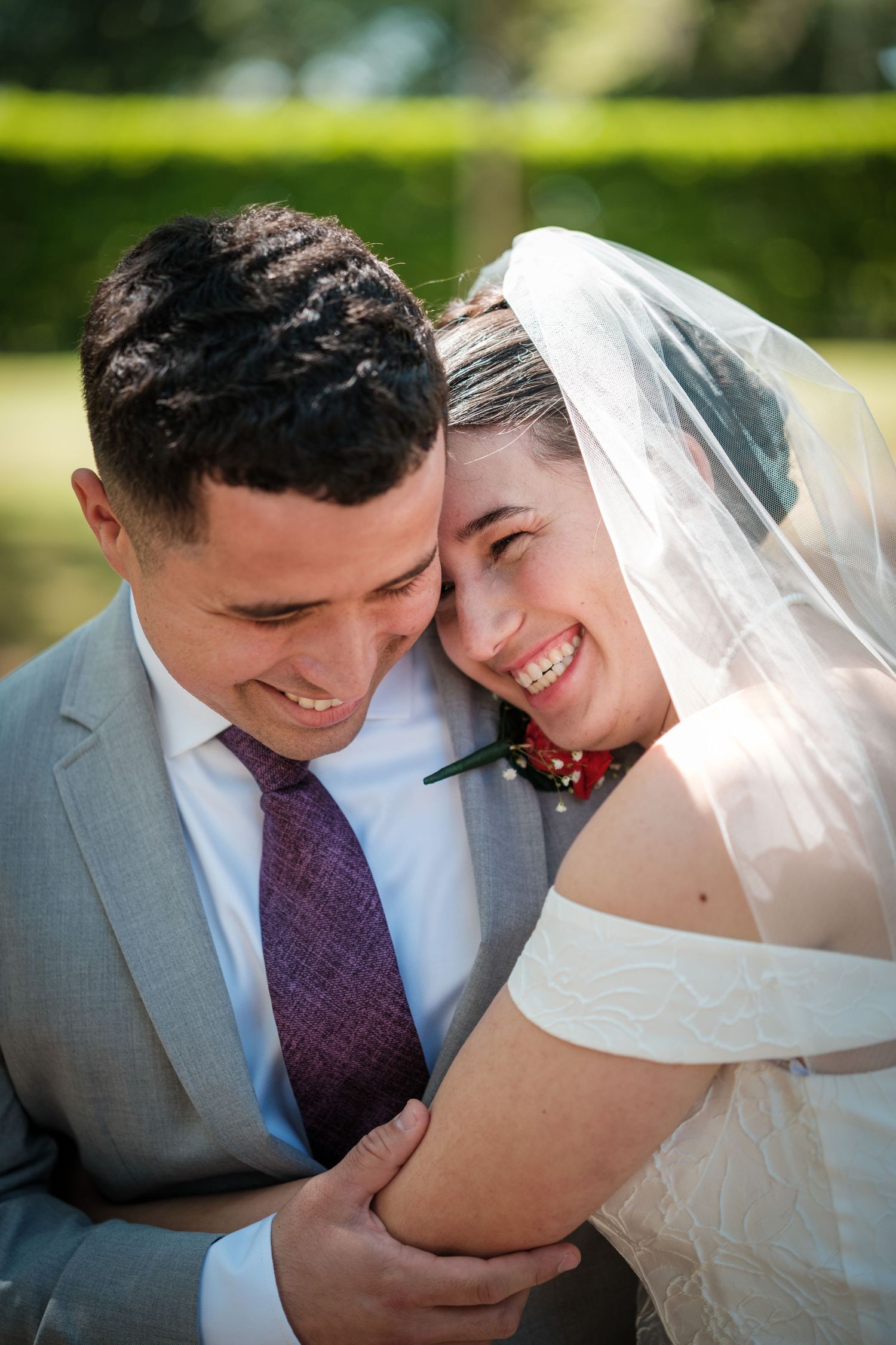 Smiling bride in white veil and groom in gray suit with purple tie share a joyful embrace outdoors.