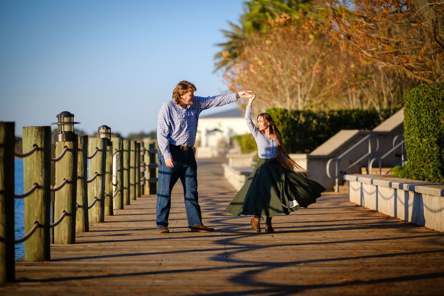 A man and young girl dance together on a wooden boardwalk near the water during golden hour.