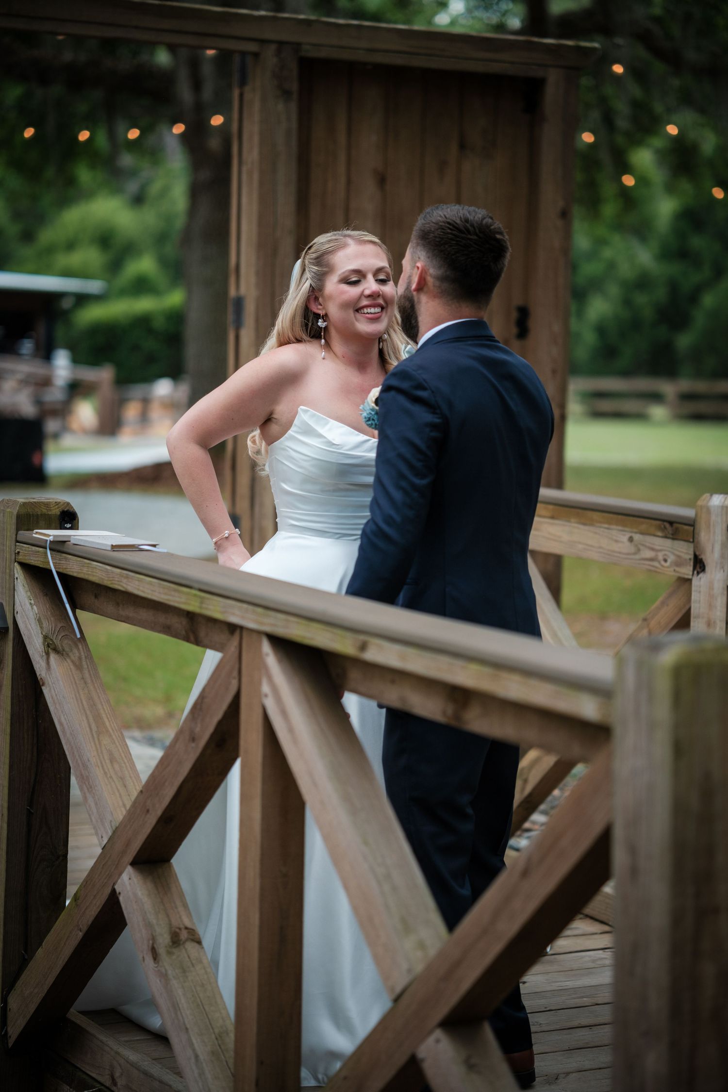 Bride in white strapless gown laughing with groom in navy suit on rustic wooden bridge at outdoor wedding venue.