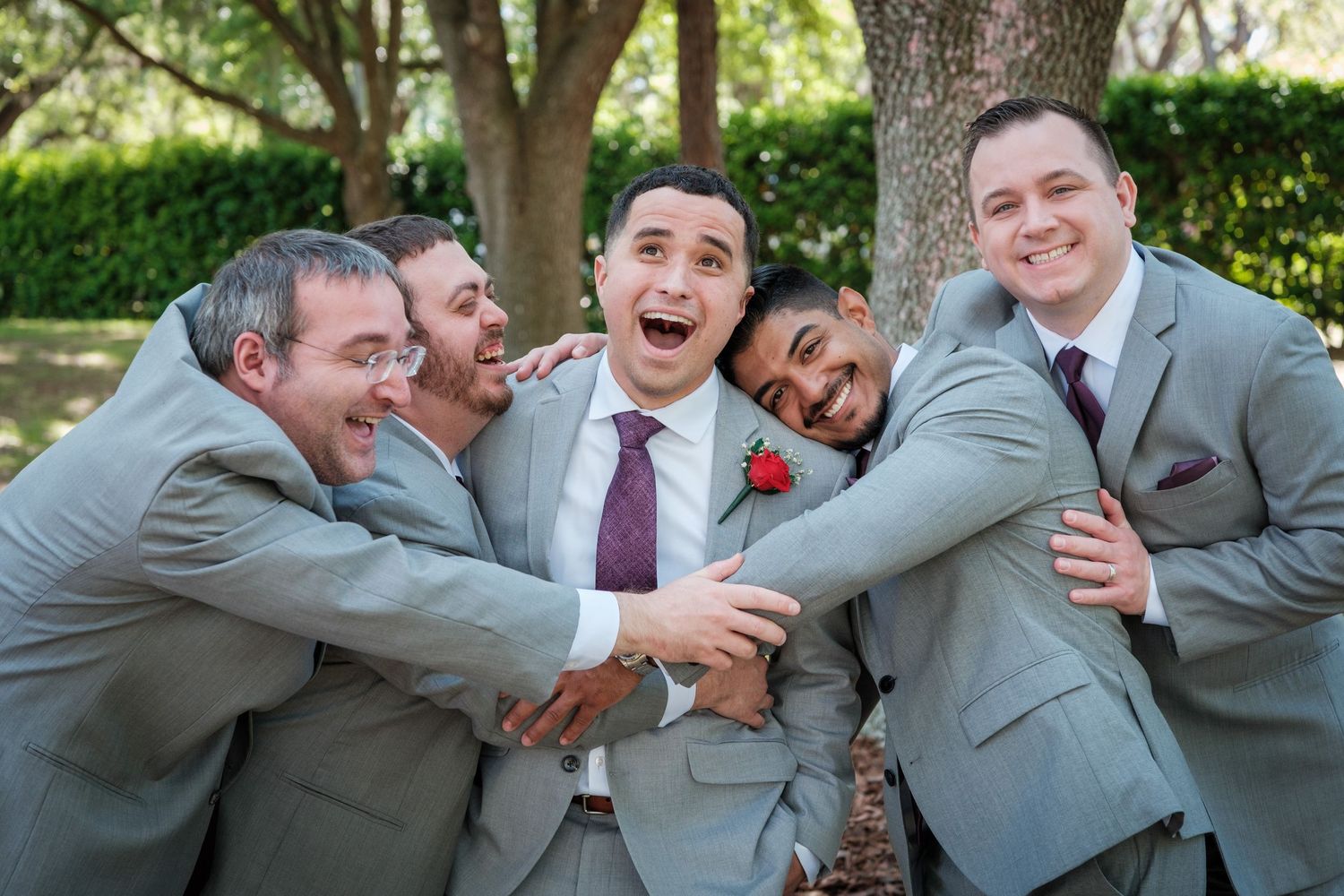Five groomsmen in gray suits celebrate joyfully outdoors, hugging and laughing together at a wedding.