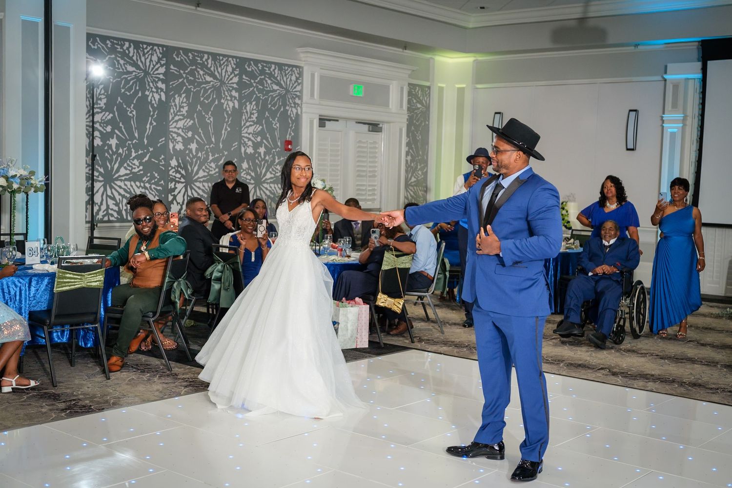 Bride and groom share first dance at elegant wedding reception with guests in blue attire watching.