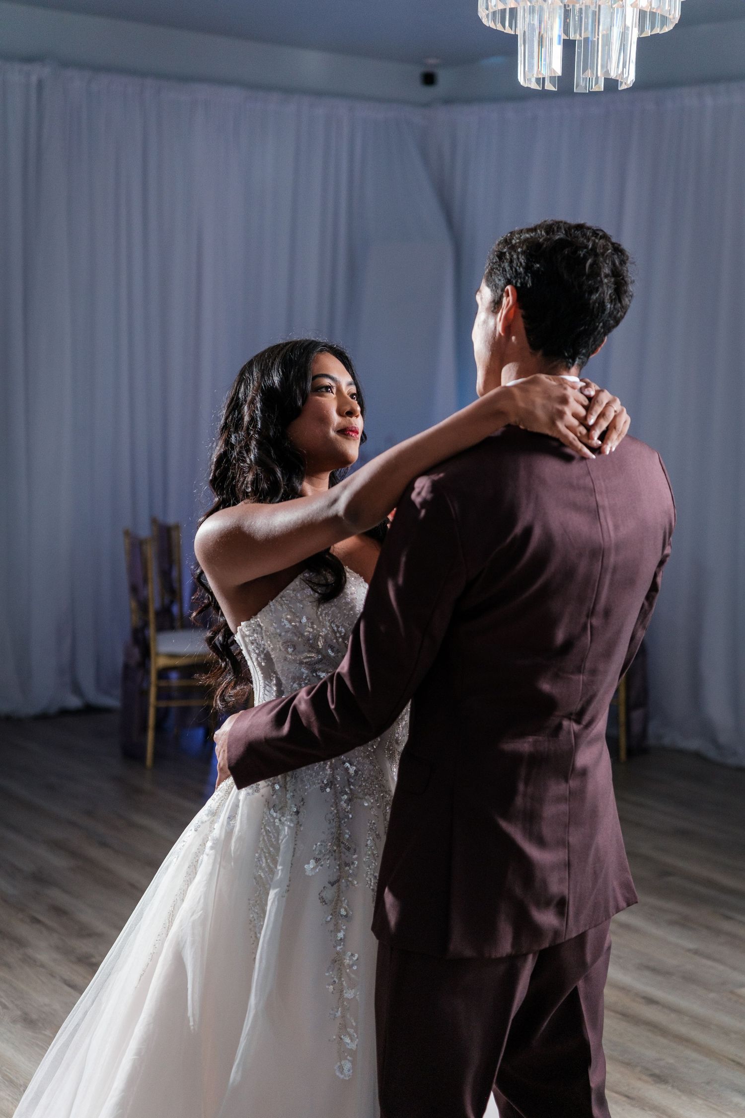 A couple shares their first dance as newlyweds in a reception hall with crystal chandeliers and white drapes.