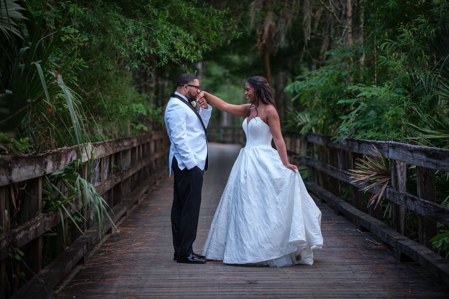 Bride and groom share a romantic moment on a wooden bridge surrounded by lush green tropical foliage.