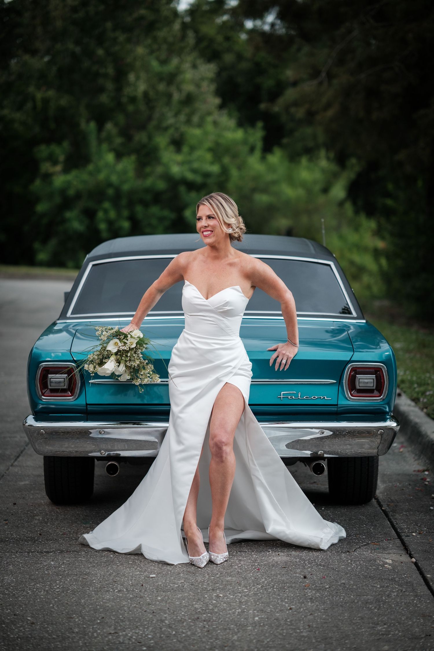 Bride in white strapless gown with slit poses against a teal classic Ford Falcon car holding a bouquet outdoors.