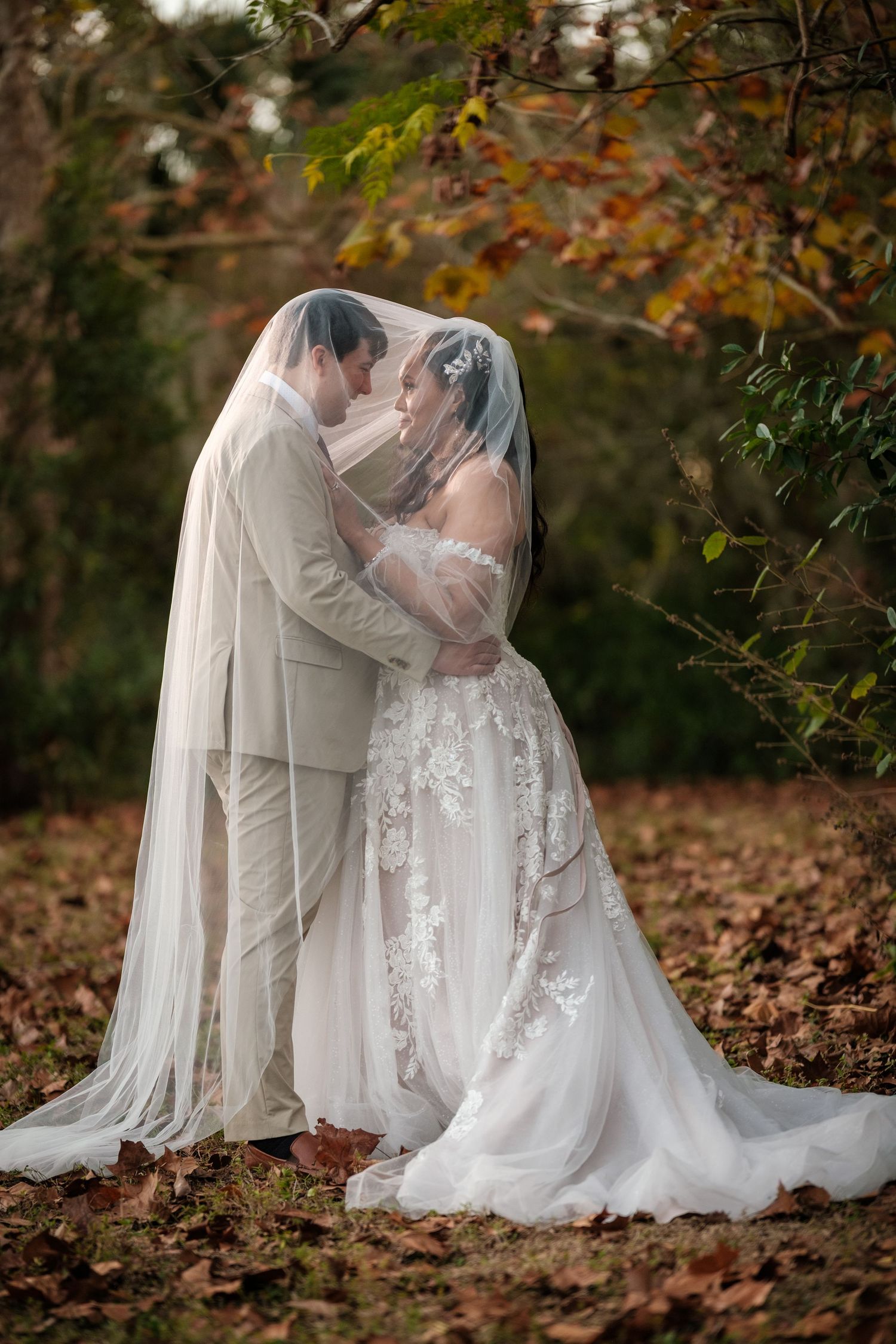 A romantic wedding couple shares an intimate moment surrounded by autumn leaves and a flowing white veil.