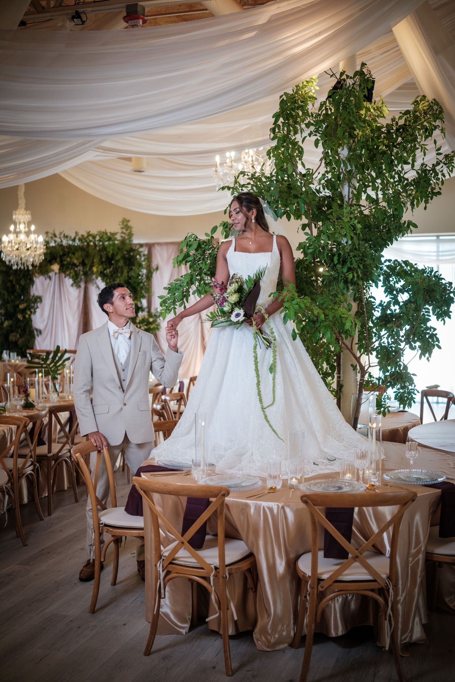 Groom gazes up at bride standing on a table surrounded by lush greenery in a beautifully decorated wedding reception hall.