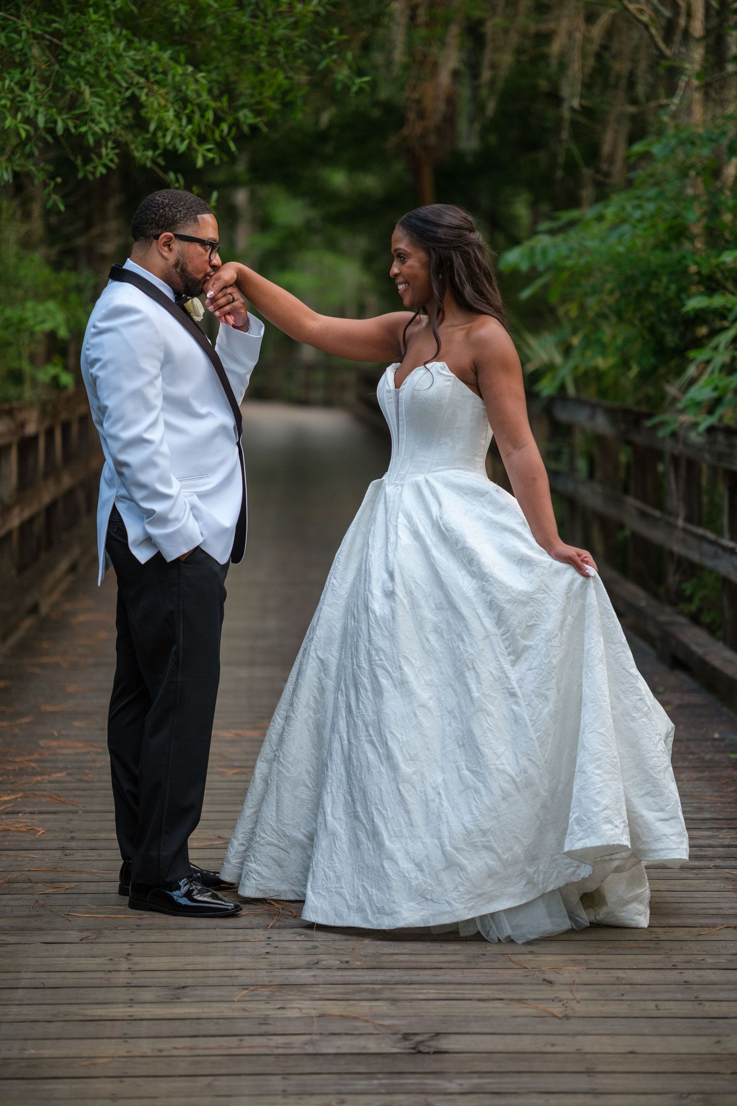 Bride and groom share a romantic hand-kiss moment on a wooden bridge surrounded by lush greenery in wedding attire.