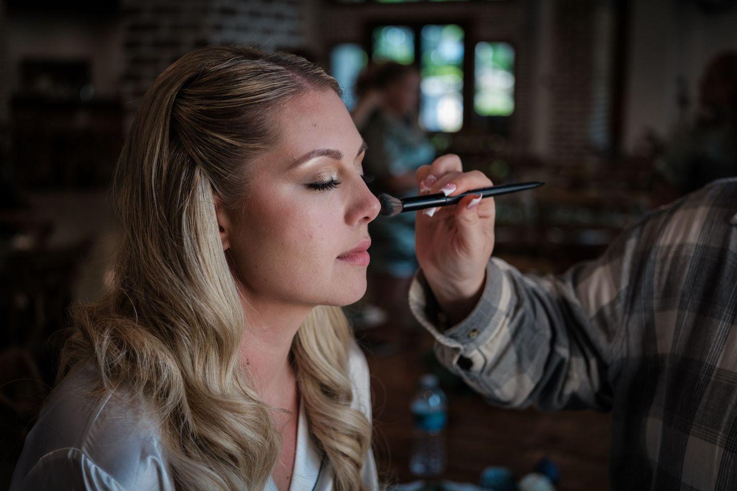 Makeup artist applying eye makeup to a blonde bride during wedding preparation in a rustic venue.