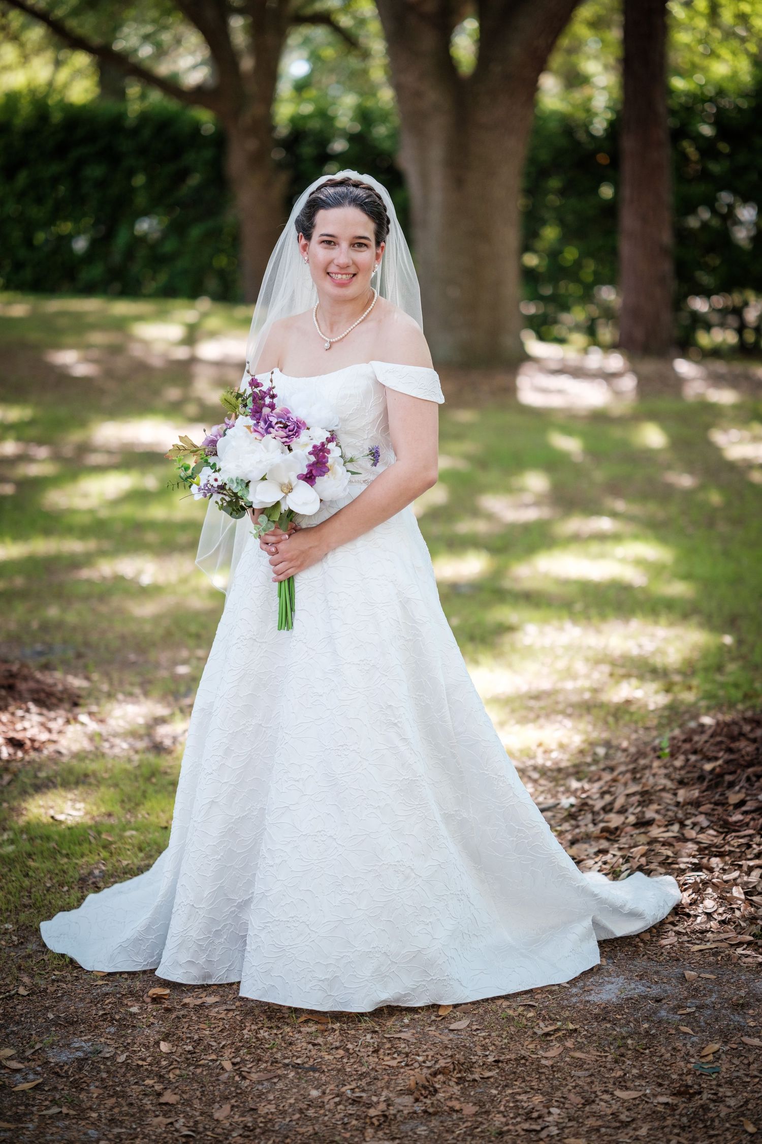 Bride in off-shoulder lace gown holding purple and white bouquet outdoors among trees.
