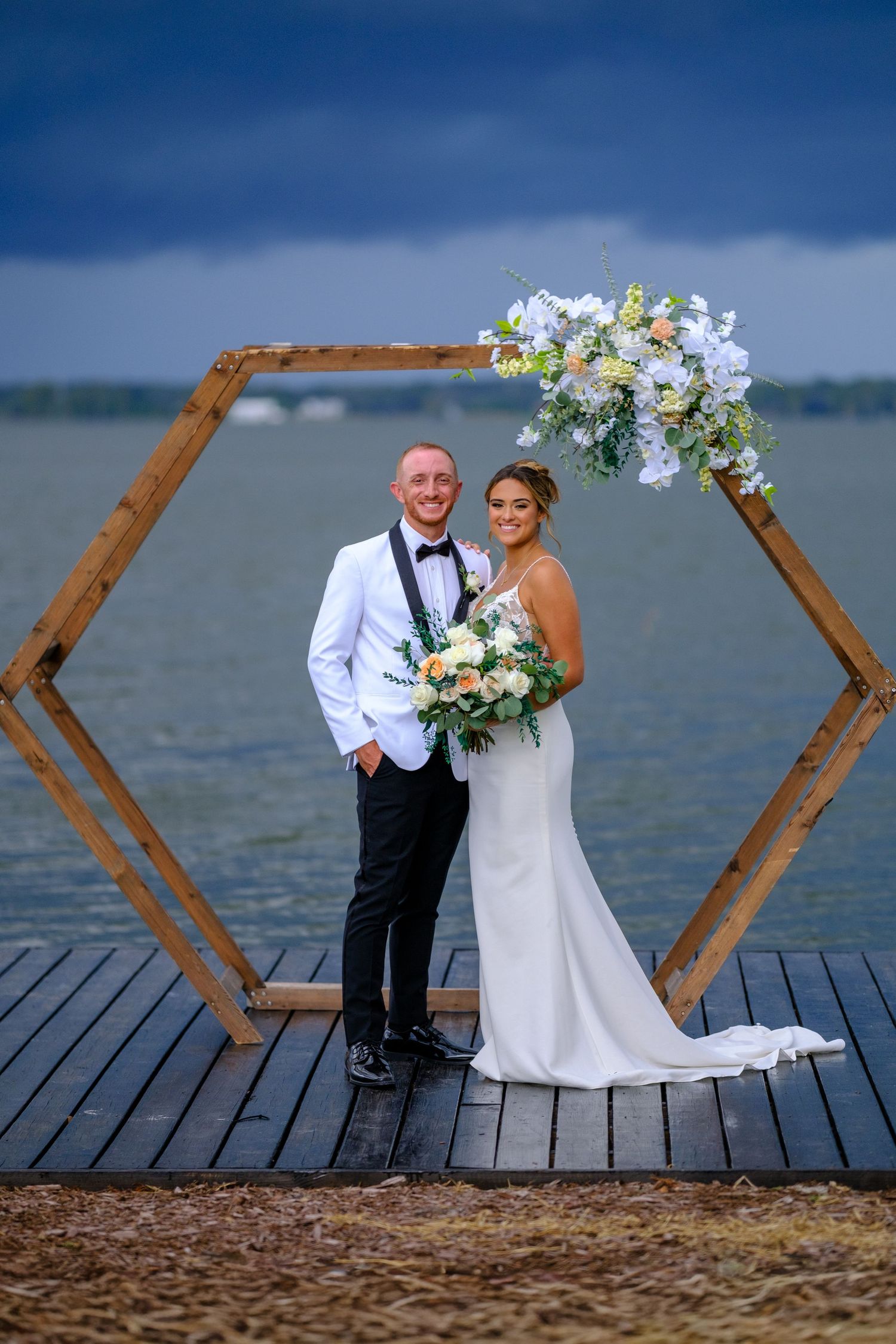 Wedding couple poses under hexagonal wooden arch decorated with white flowers on waterfront deck during stormy day.