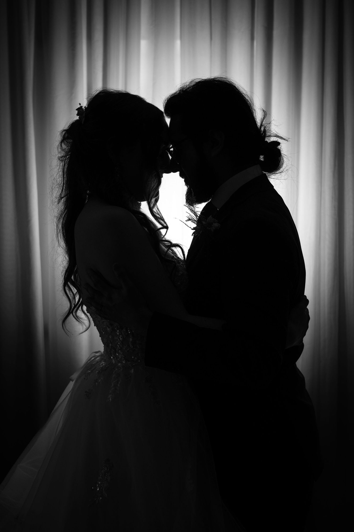 Black and white silhouette of bride and groom touching foreheads romantically against a curtain backdrop.