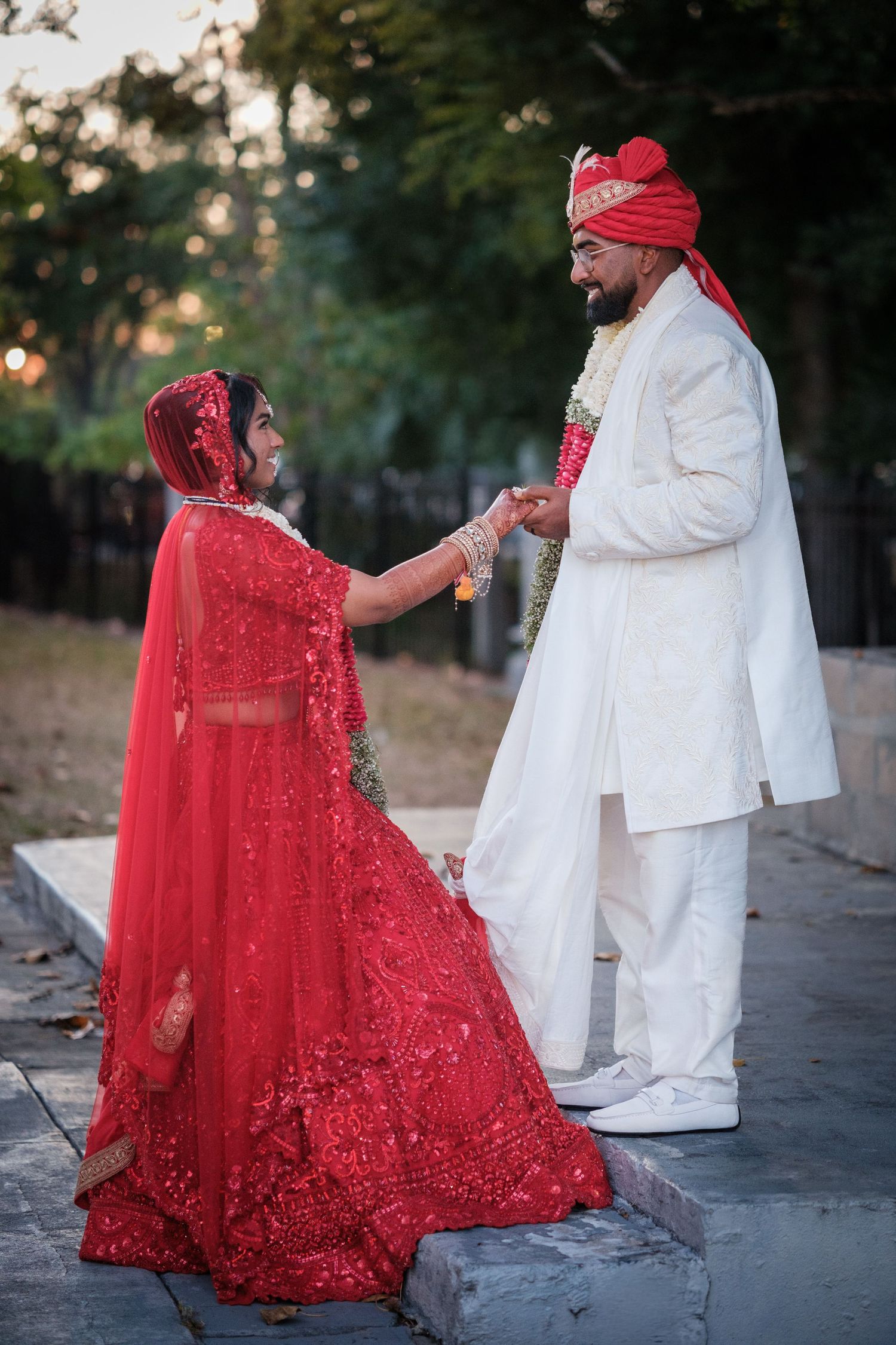 Indian bride in red lehenga and groom in white sherwani holding hands during traditional wedding ceremony outdoors.