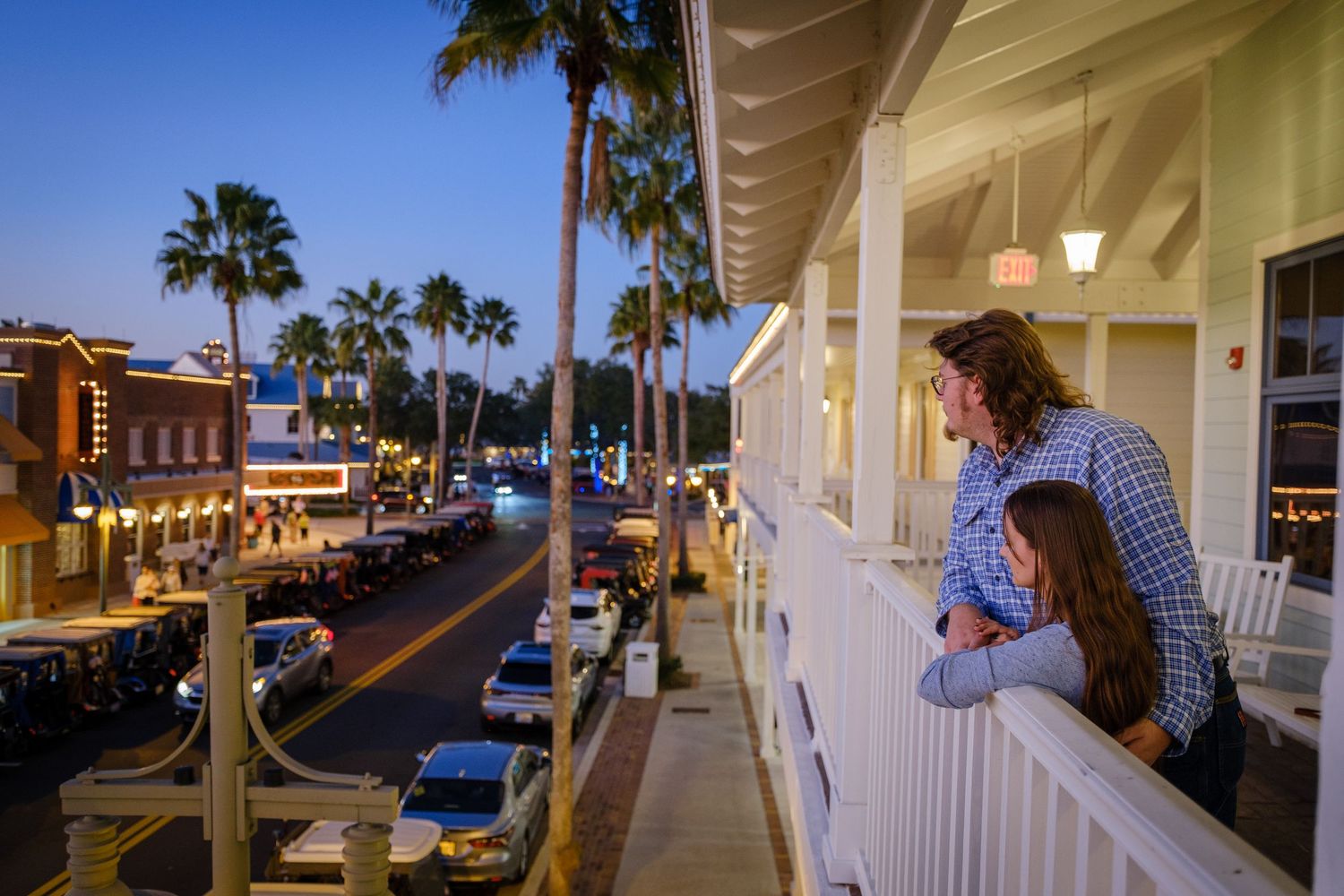 Couple standing on a balcony overlooking a charming lit street lined with palm trees at dusk.