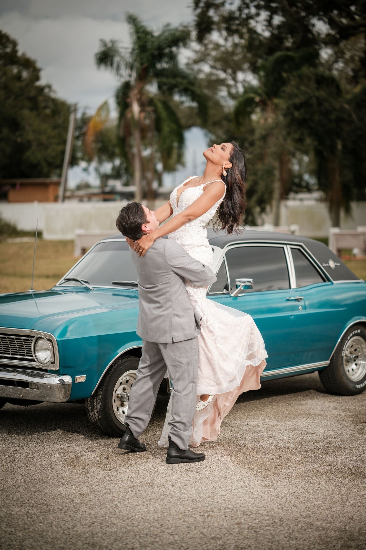 Wedding couple poses playfully near a vintage turquoise car on a gravel surface.