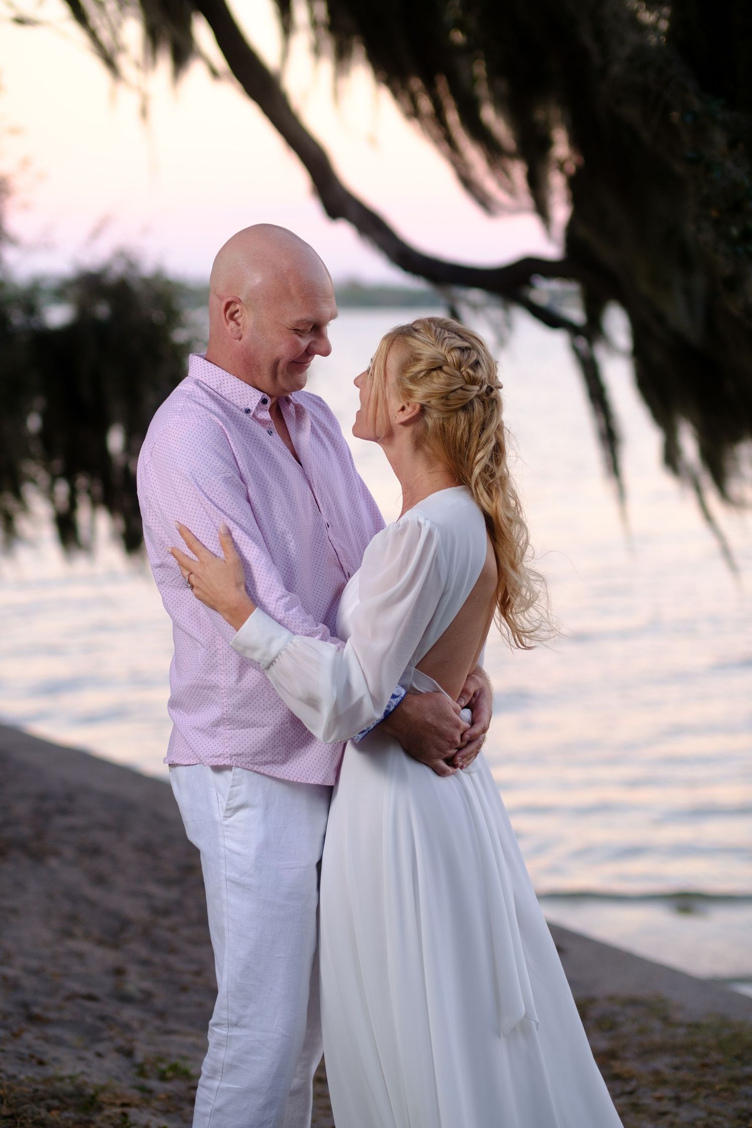 A romantic couple in white and lavender attire embraces on a beach at sunset with Spanish moss trees overhead.