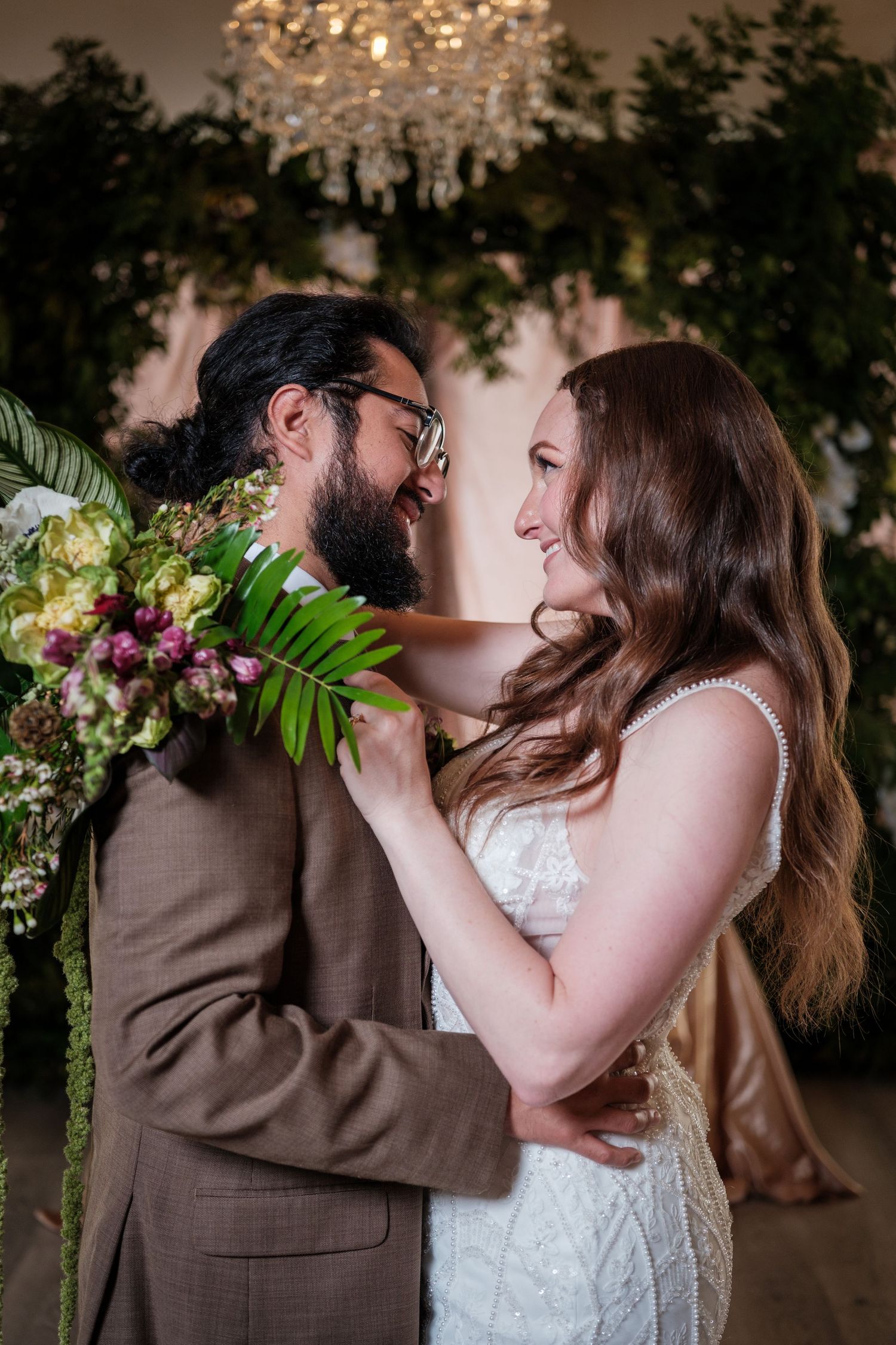 A bearded groom in a brown suit and bride in a lace dress share a romantic moment surrounded by lush greenery and flowers.