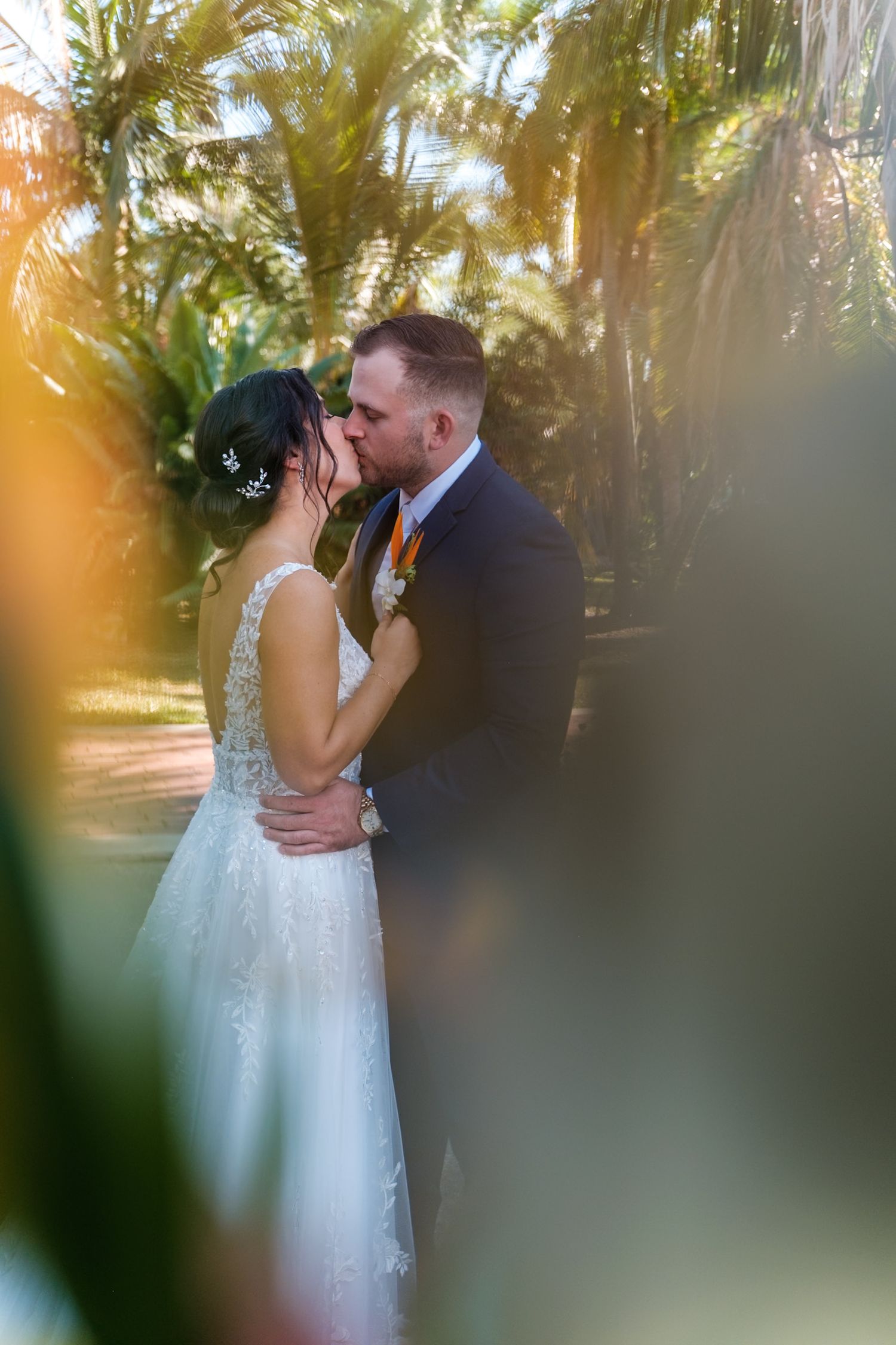 A romantic wedding couple shares a tender kiss outdoors with palm trees and sunlight creating a dreamy atmosphere.