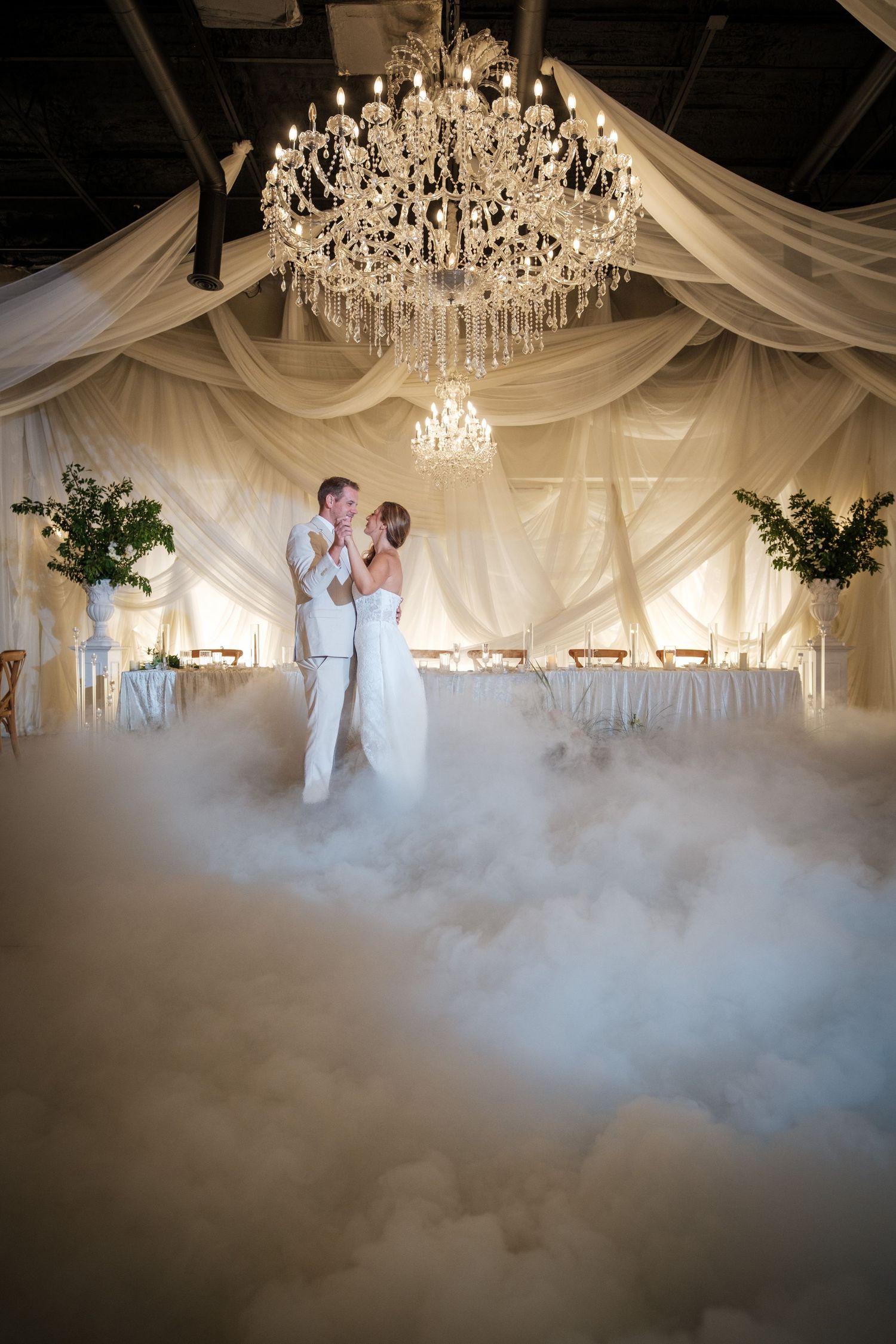 Bride and groom share first dance surrounded by dramatic fog under crystal chandeliers in elegant white draped wedding venue.