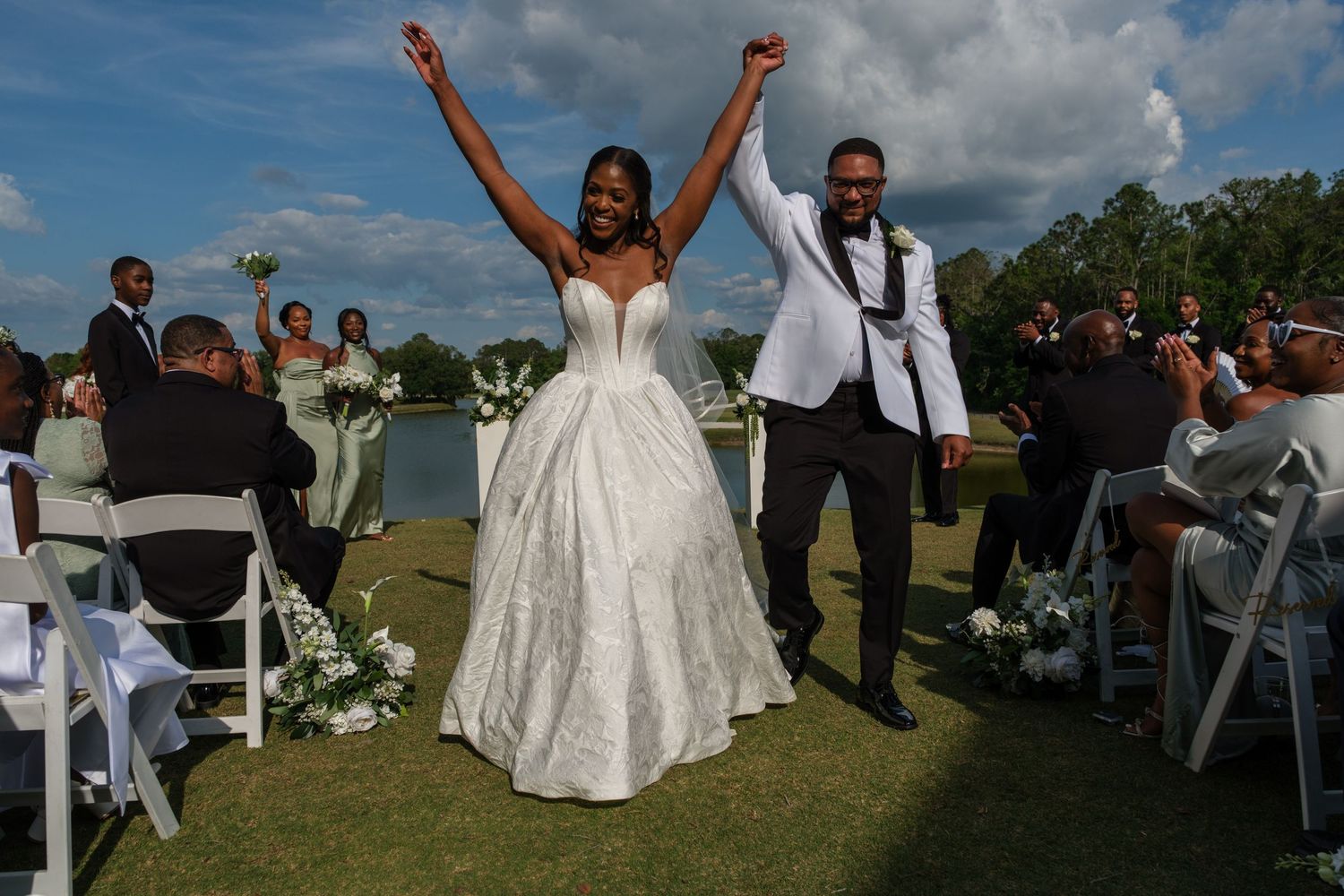 Joyful newlyweds celebrate after outdoor wedding ceremony by lakeside, bride in white gown raises arms in triumph.