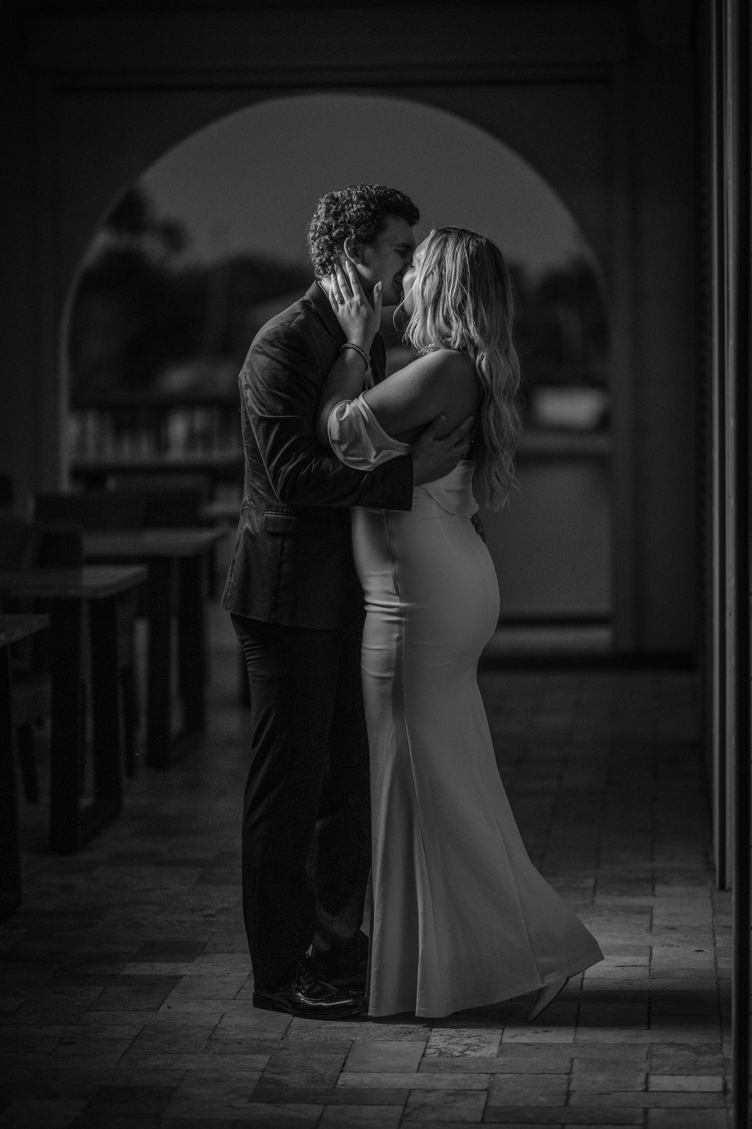 Black and white photo of a couple sharing a romantic kiss in an elegant arched hallway.