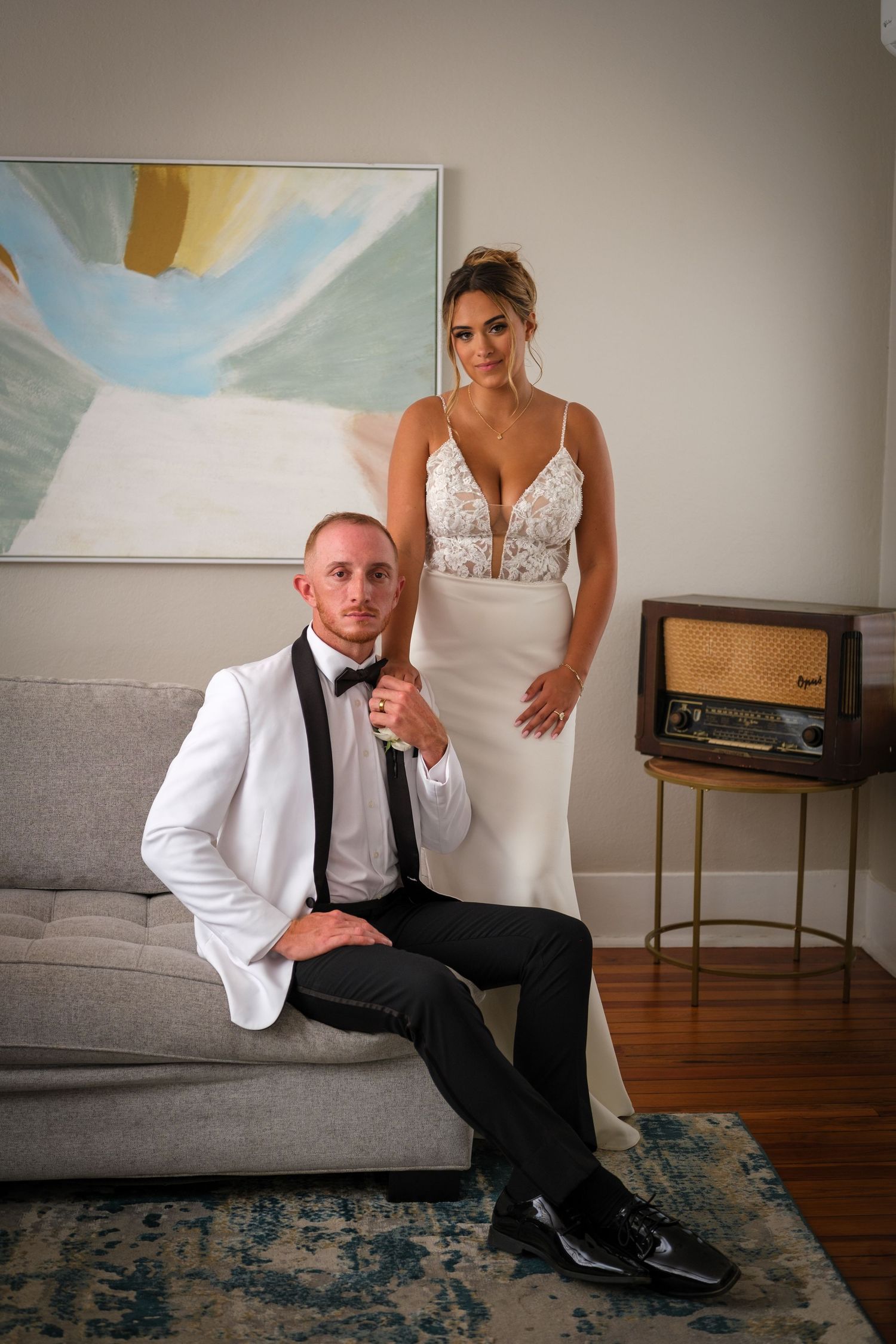 Elegant couple in formal attire pose in living room with vintage radio and abstract art on wall.