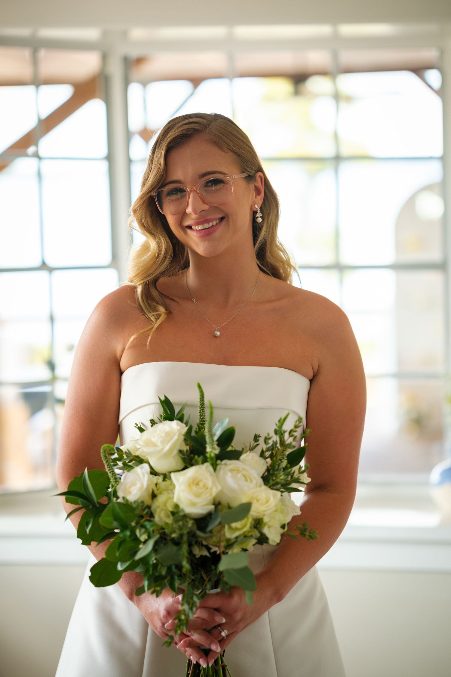 Bride in strapless white dress holds elegant bouquet of white roses and greenery while smiling indoors.