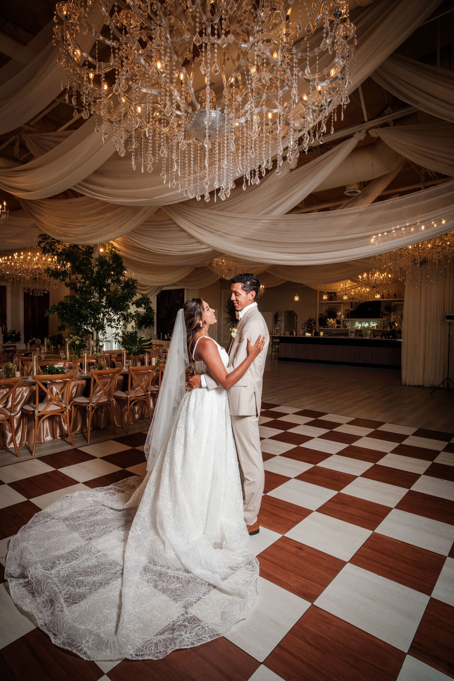 Bride and groom share a tender embrace on a checkered dance floor beneath a grand crystal chandelier.