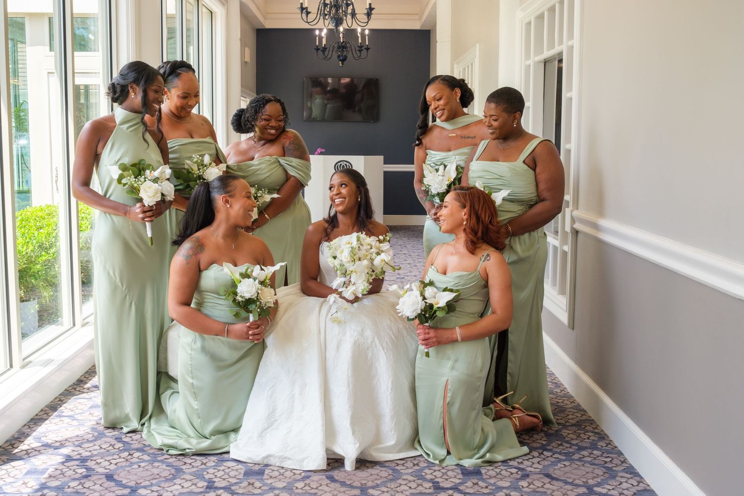 Bride in white gown surrounded by six bridesmaids in sage green dresses holding white bouquets in elegant venue hallway.