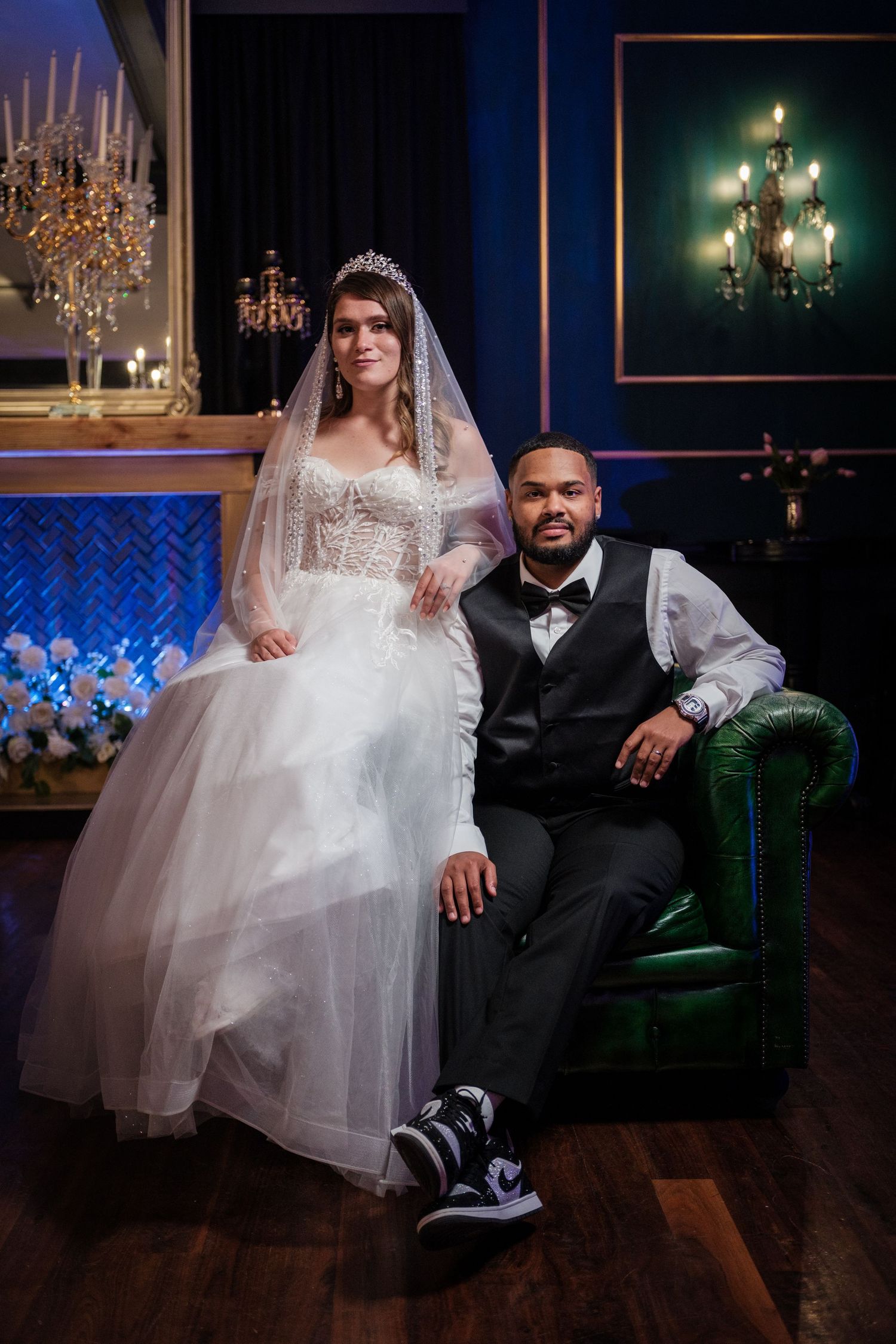 Bride in white gown and groom in black suit pose elegantly in a luxurious blue room with chandeliers.