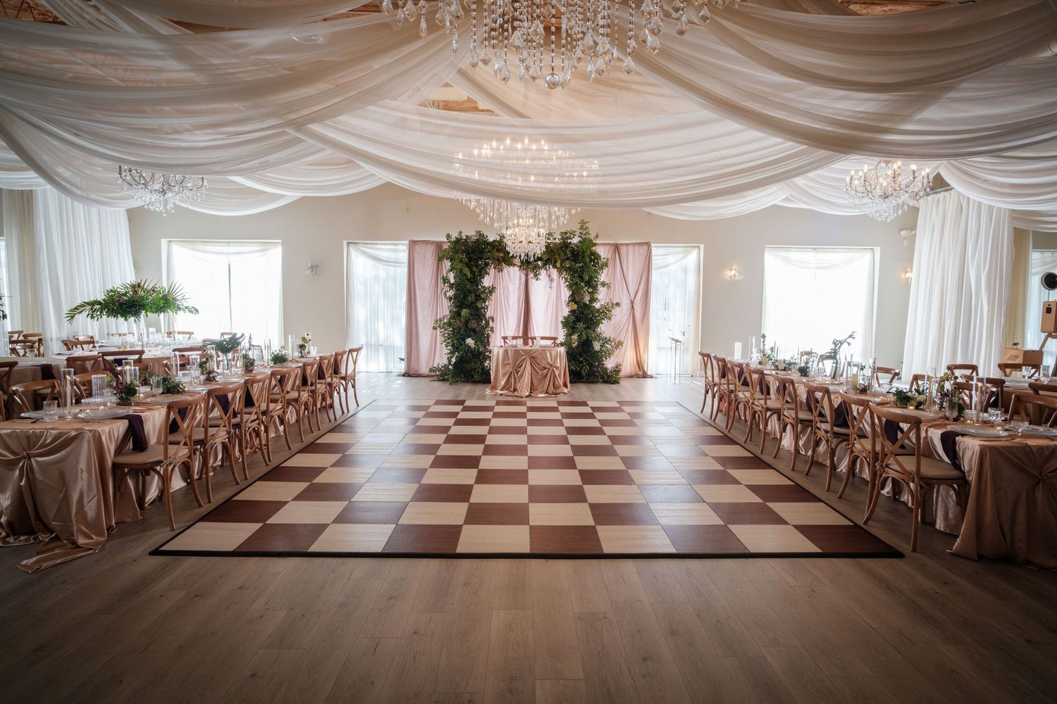 Elegant wedding reception hall with checkered dance floor, draped white ceiling, chandeliers, and rose gold table settings.