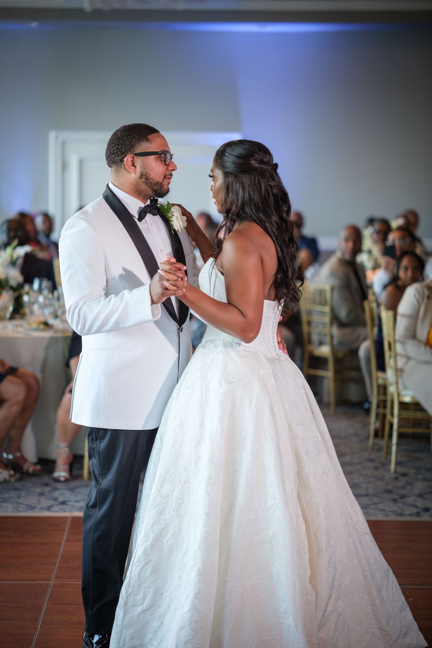 Bride and groom share first dance in ballroom, groom in white tuxedo jacket with bow tie.
