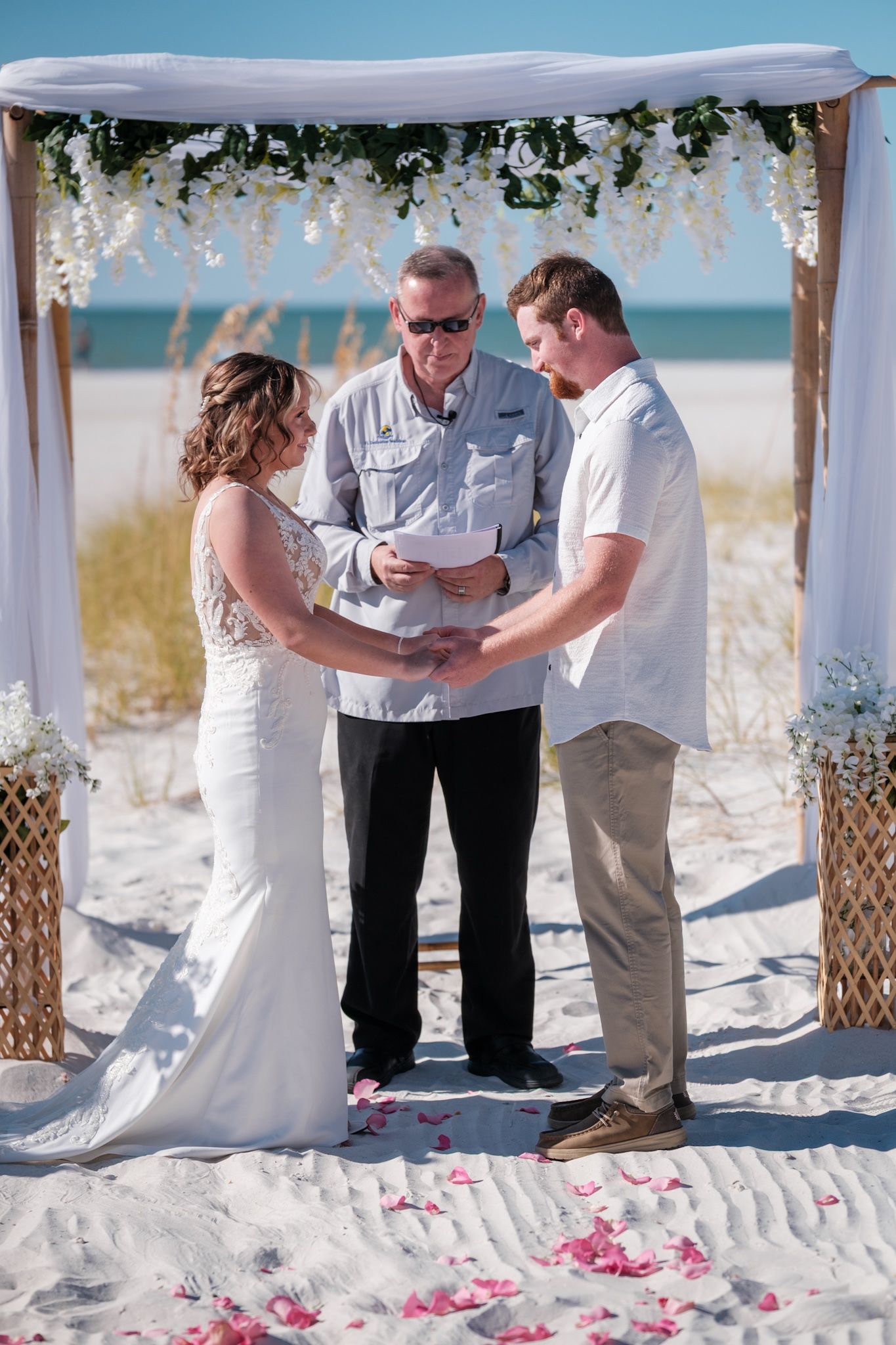 A beach wedding ceremony under a white canopy decorated with flowers and greenery on a sunny day.