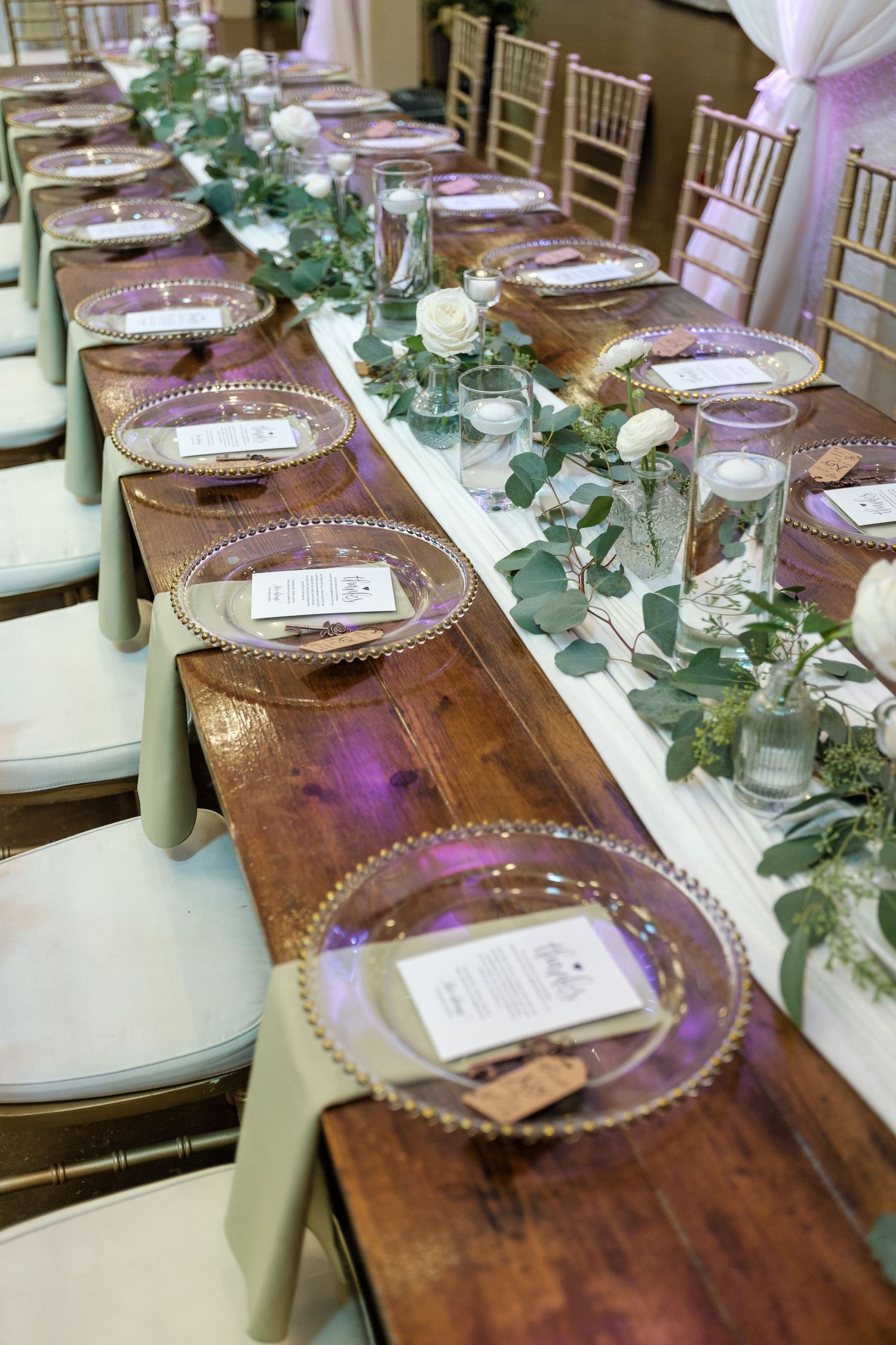 Rustic wooden banquet table set with elegant glass charger plates and eucalyptus garland for a wedding reception.
