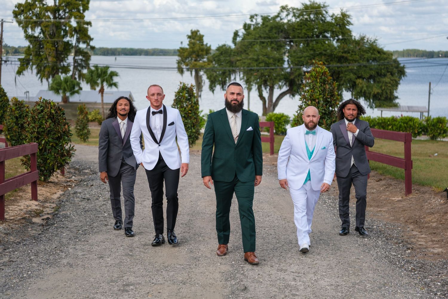 Wedding party walking together along waterfront path with trees and lake in background.