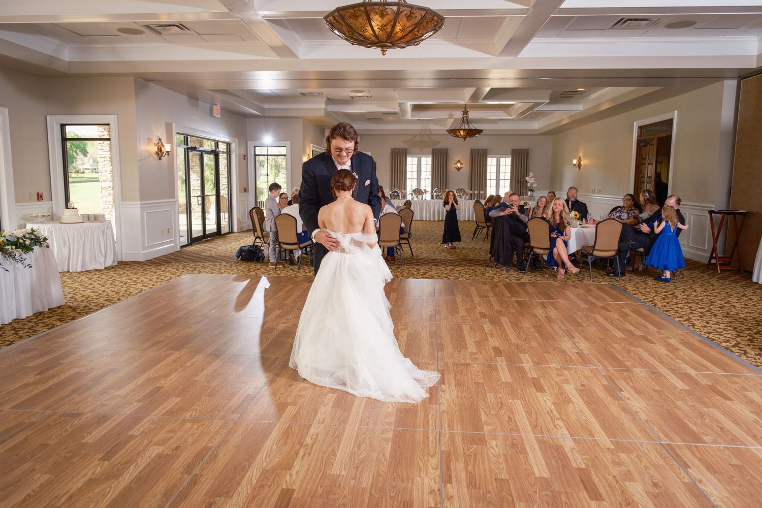 Bride and groom share their first dance on a wooden floor in an elegant ballroom with guests seated around them.
